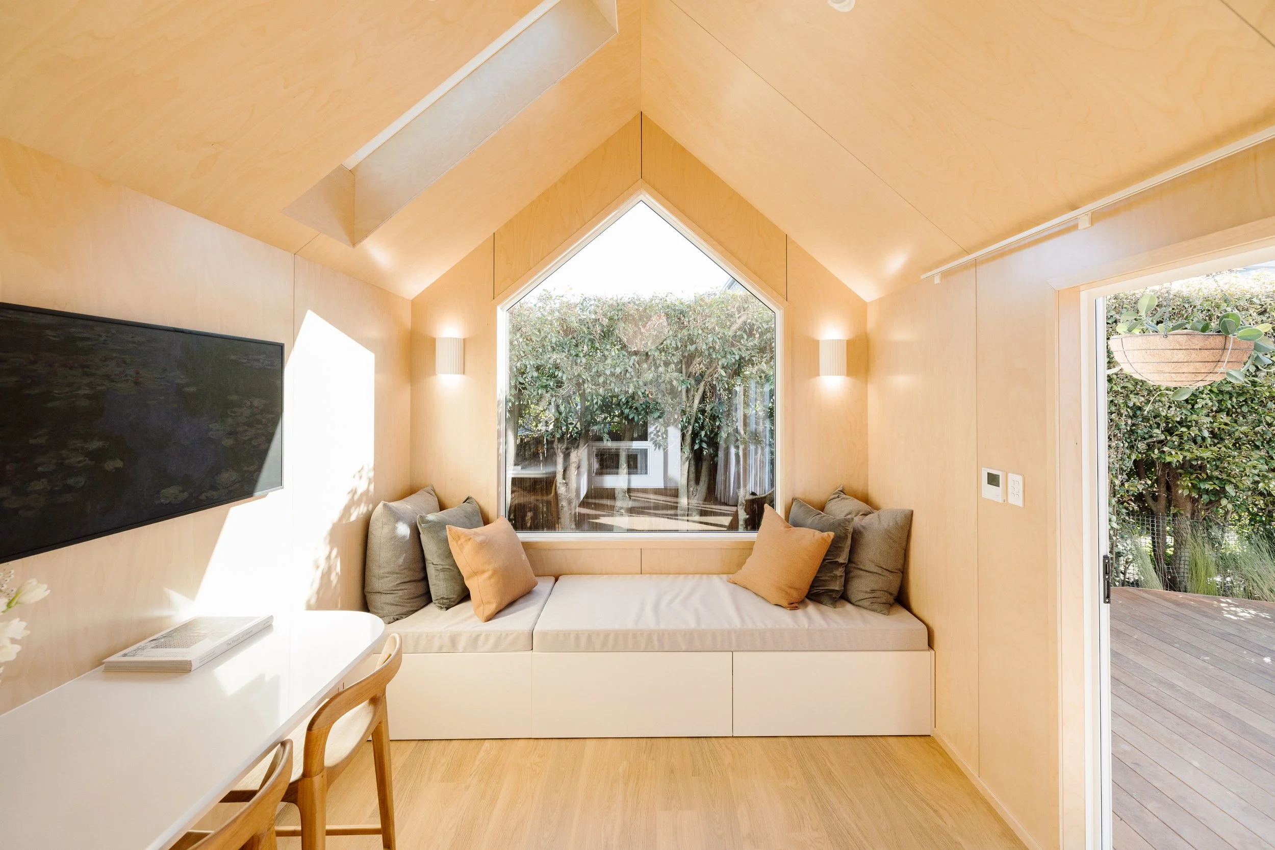 Bright living room with wood-paneled walls and ceiling, featuring a large front window with trees outside, a cushioned bench with pillows, a wall-mounted TV, and a sliding glass door leading to a patio.