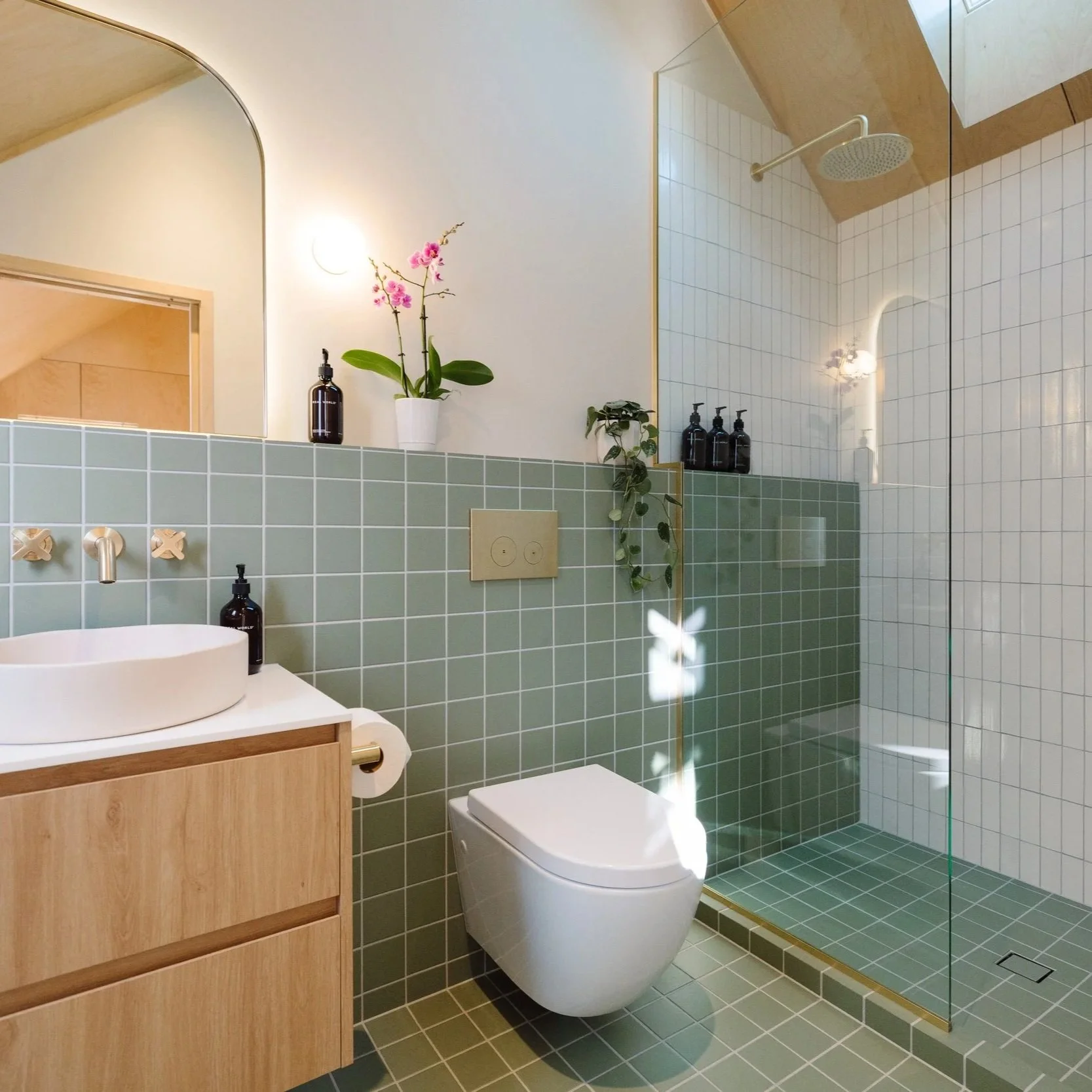 A modern bathroom with green tiled walls and floor, a white circular vessel sink on a wooden vanity, a toilet, a glass shower with a tiled interior and a rainfall showerhead, a potted orchid and a leafy plant on a ledge, and a large mirror above the sink reflecting the shower area.