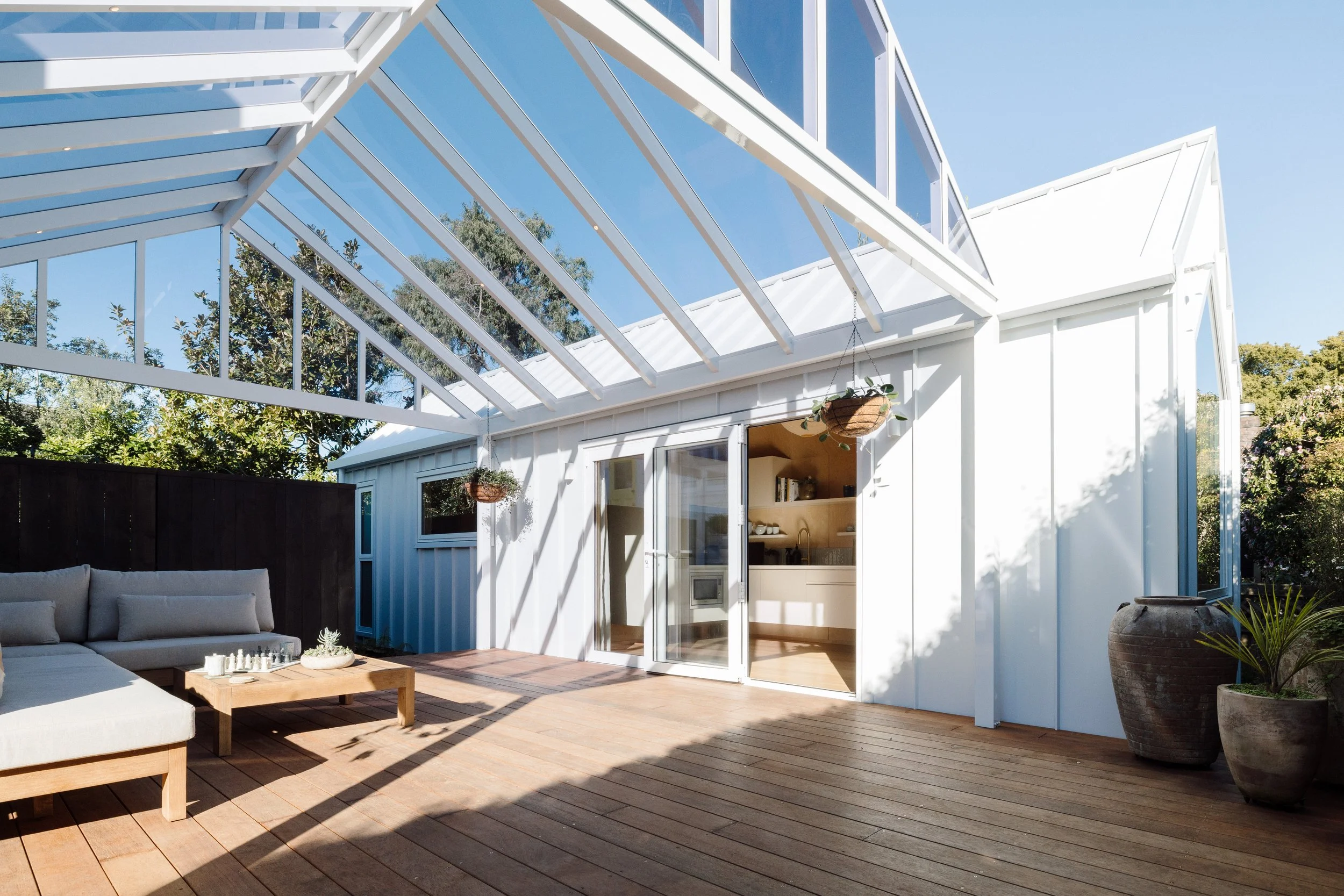 A modern outdoor patio area with a wooden deck, a white outdoor sofa with cushions, a coffee table with a chess set, and hanging plants. An adjacent white building features large glass doors and windows. A glass roof structure extends from the building, with trees visible in the background under a clear blue sky.