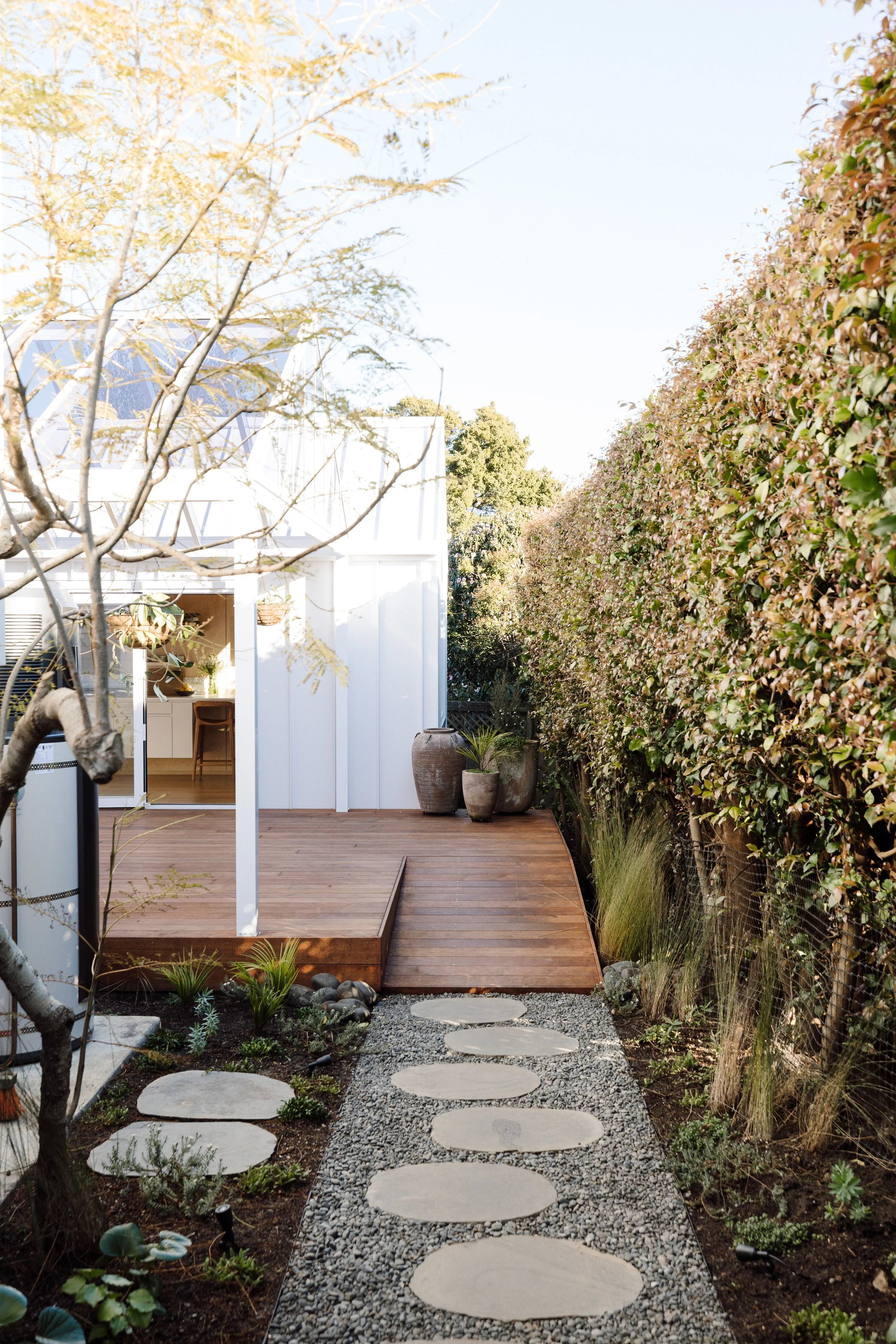 A backyard garden with a circular stepping stone pathway on gravel, bordered by various plants and shrubs, leading to a wooden deck attached to a modern white house with an open door revealing a kitchen interior.