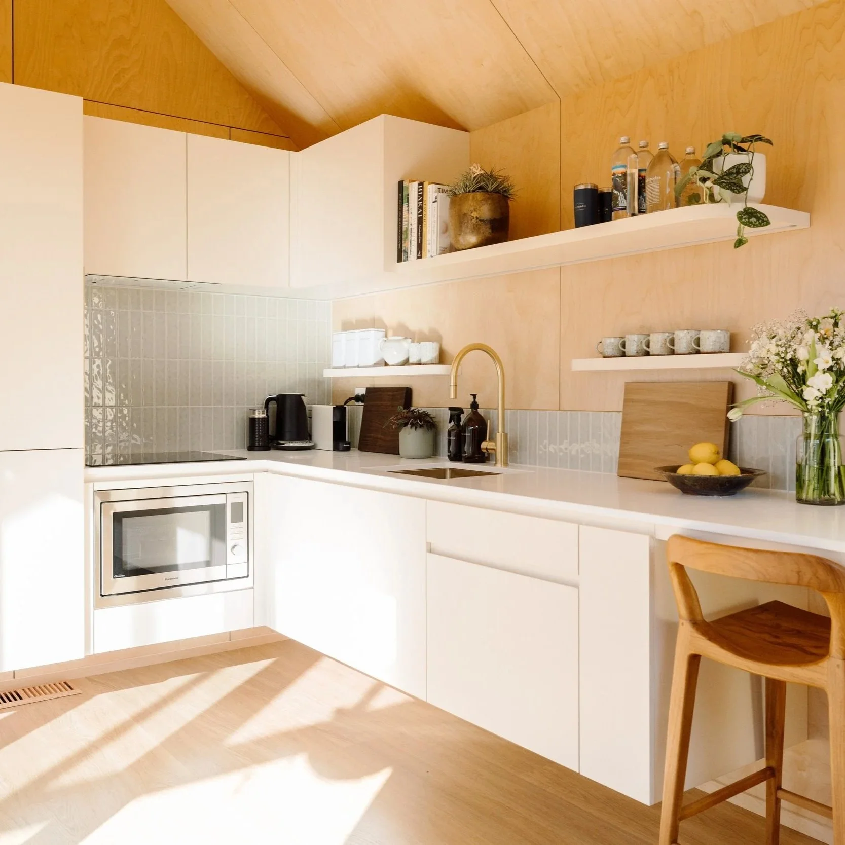 Modern full kitchen with white cabinets, a stone countertop, and open shelves decorated with books, potted plants, and drinking water bottles. A oven and cooktop is built into the cabinetry, and a wooden chair is partially visible in the foreground.