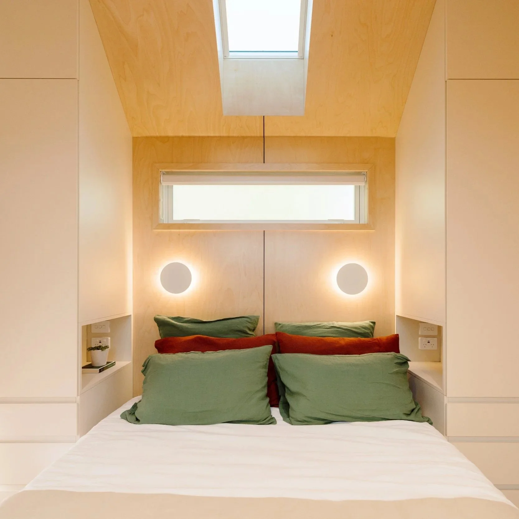 A bedroom with a bed featuring green and pillows, birch plywood walls with a window and skylight above, and built-in side shelves with plants and books.