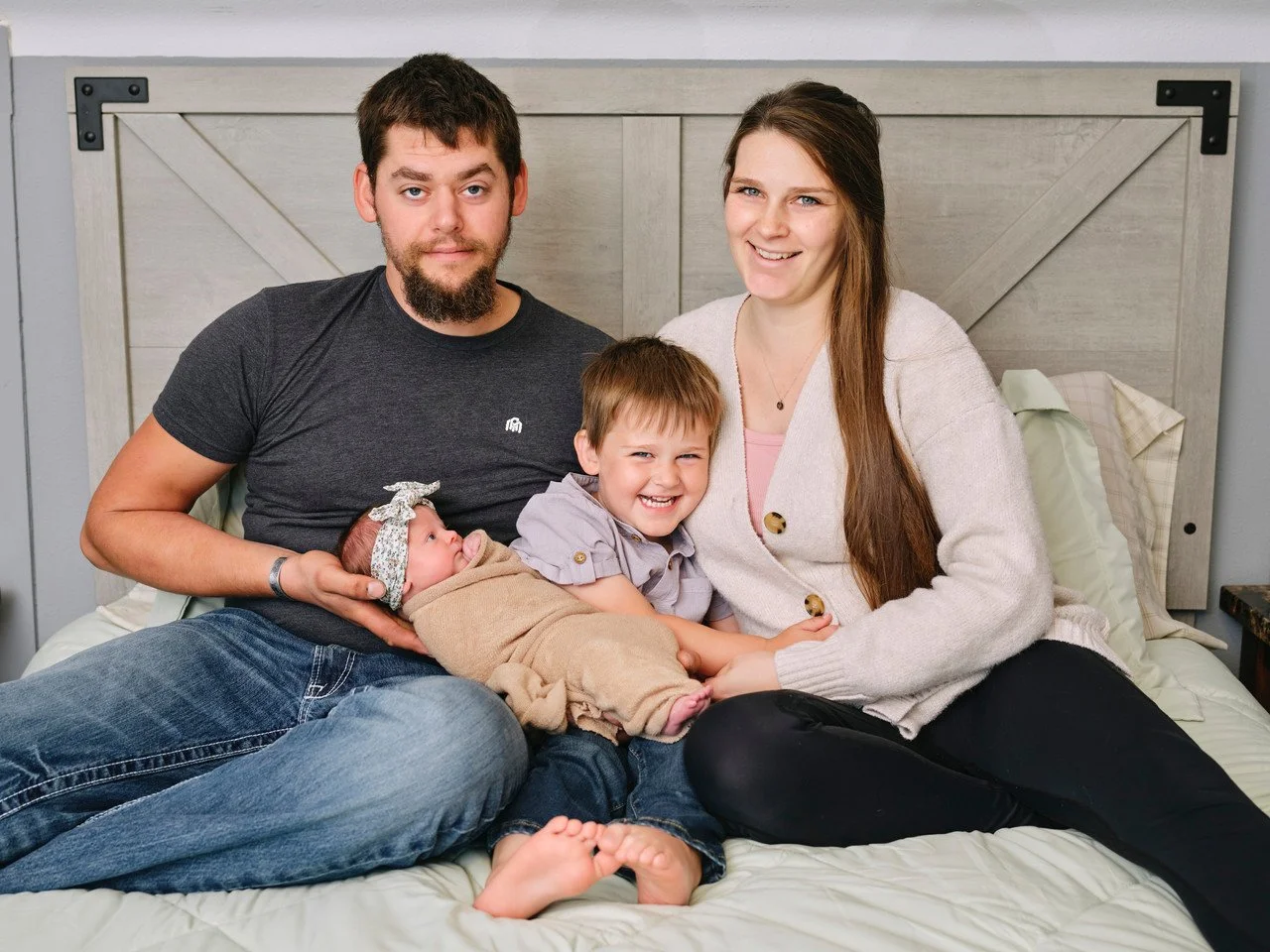 Family of four sitting on a bed, including a man, woman, young boy, and a newborn girl, smiling and enjoying family time.