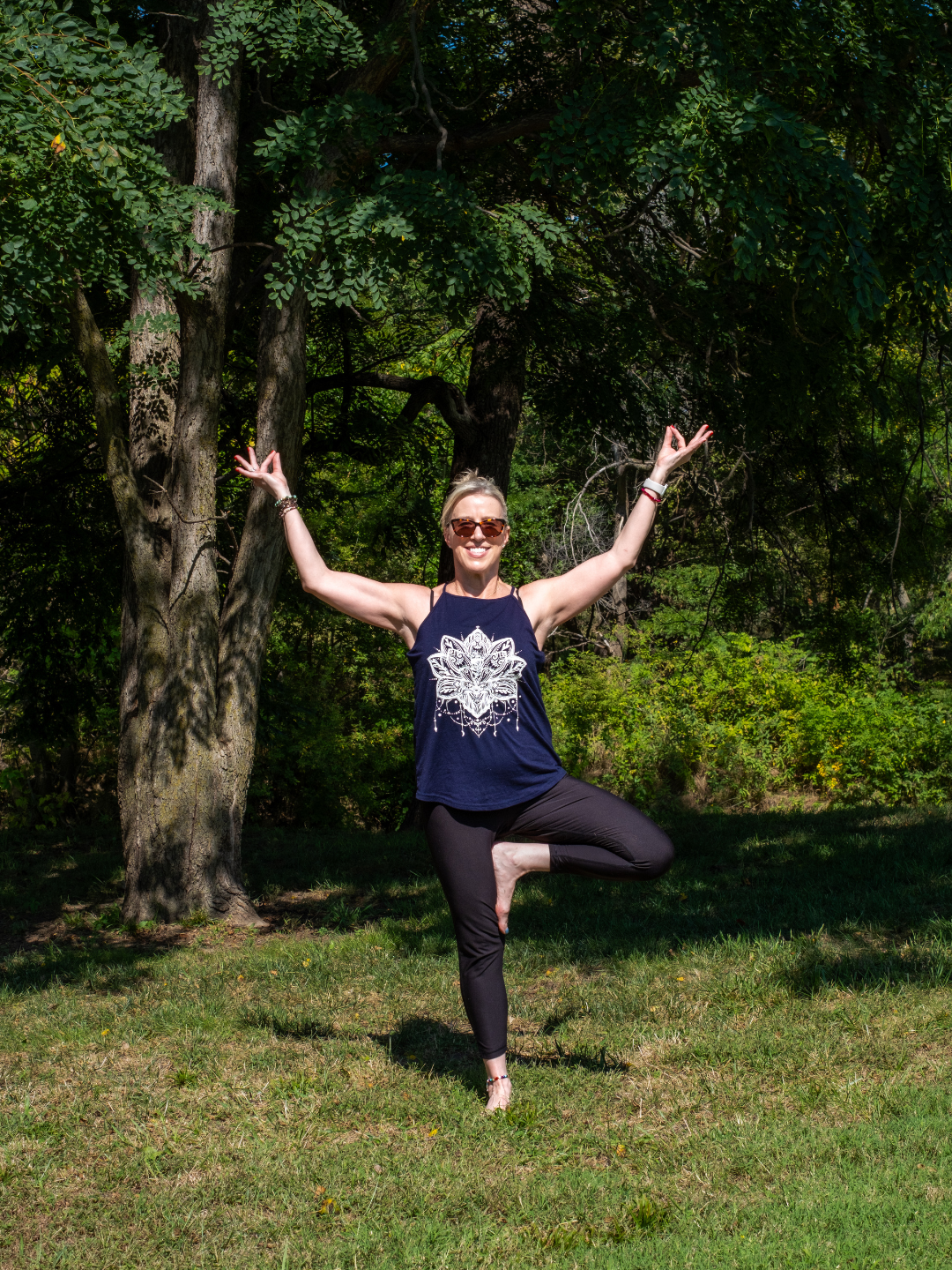 Woman in yoga pose outdoors, standing on grass in front of trees, wearing sunglasses, a blue tank top with a white wolf design, and black leggings.