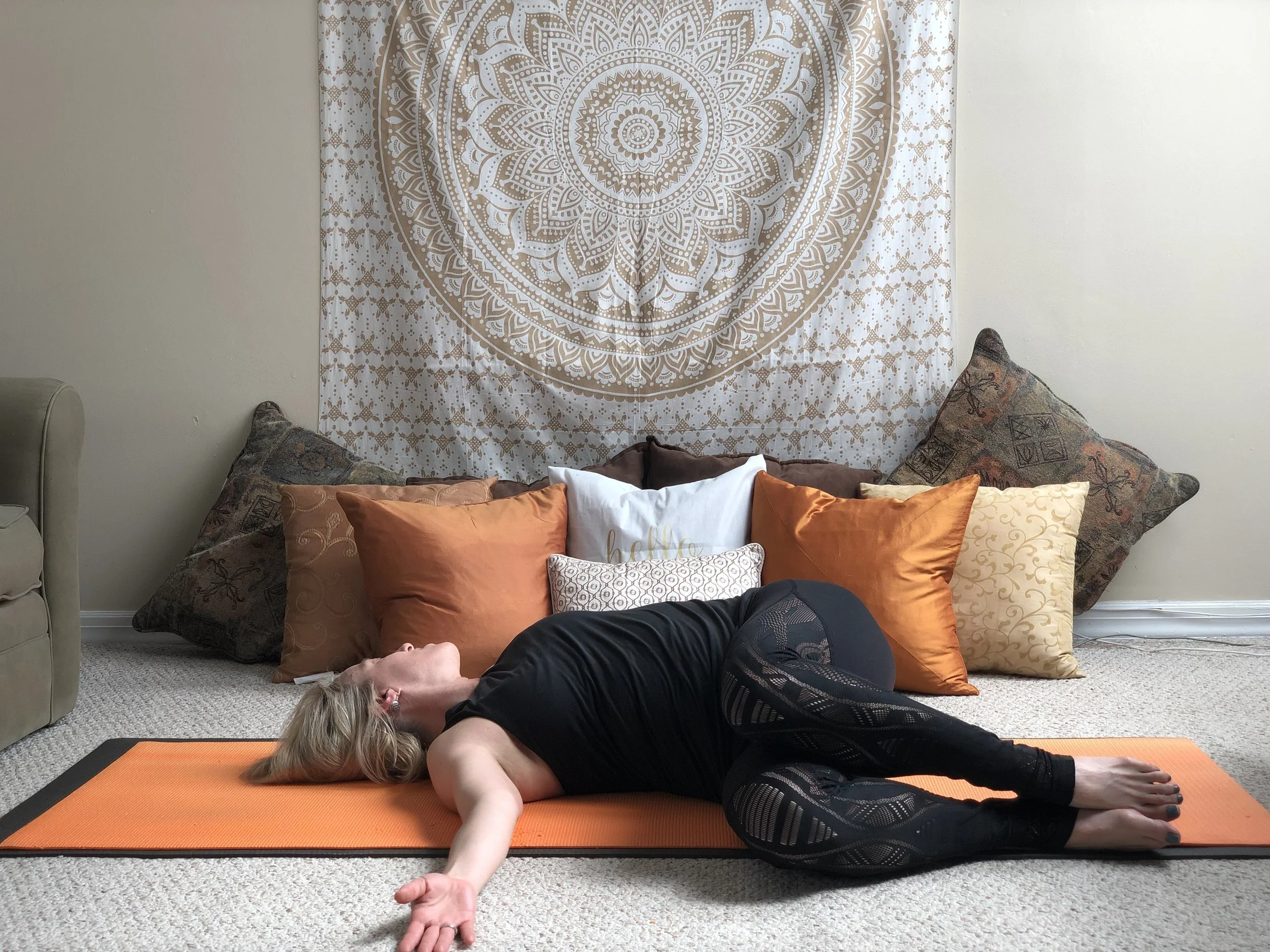 A woman practicing yoga on an orange mat in a room decorated with a large, intricate circular mandala tapestry hanging on the wall. She is lying on her side with her eyes closed, wearing black athletic clothing, and surrounded by multiple pillows.