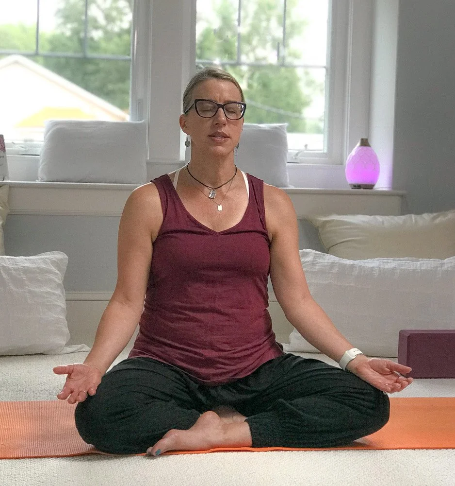 A woman practicing yoga in a bright living room, sitting cross-legged with hands on her knees in a meditative pose, wearing glasses and a maroon tank top.