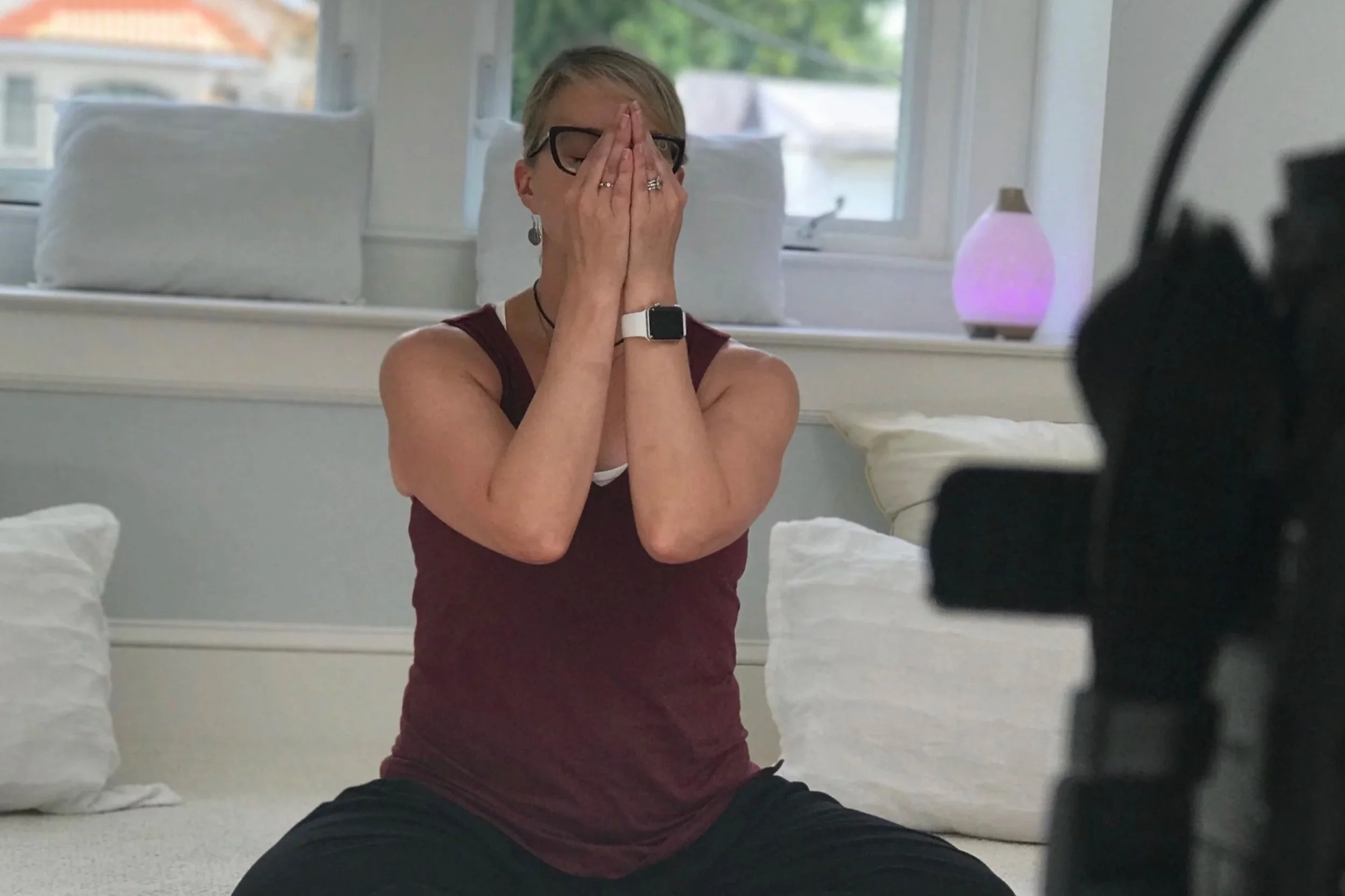 A woman wearing glasses and a maroon tank top sitting on a bed with white pillows, holding her hands together in front of her face in a prayer position, with a yoga mat and a black dog near her. There is a window with a view of trees and a Himalayan salt lamp in the background.