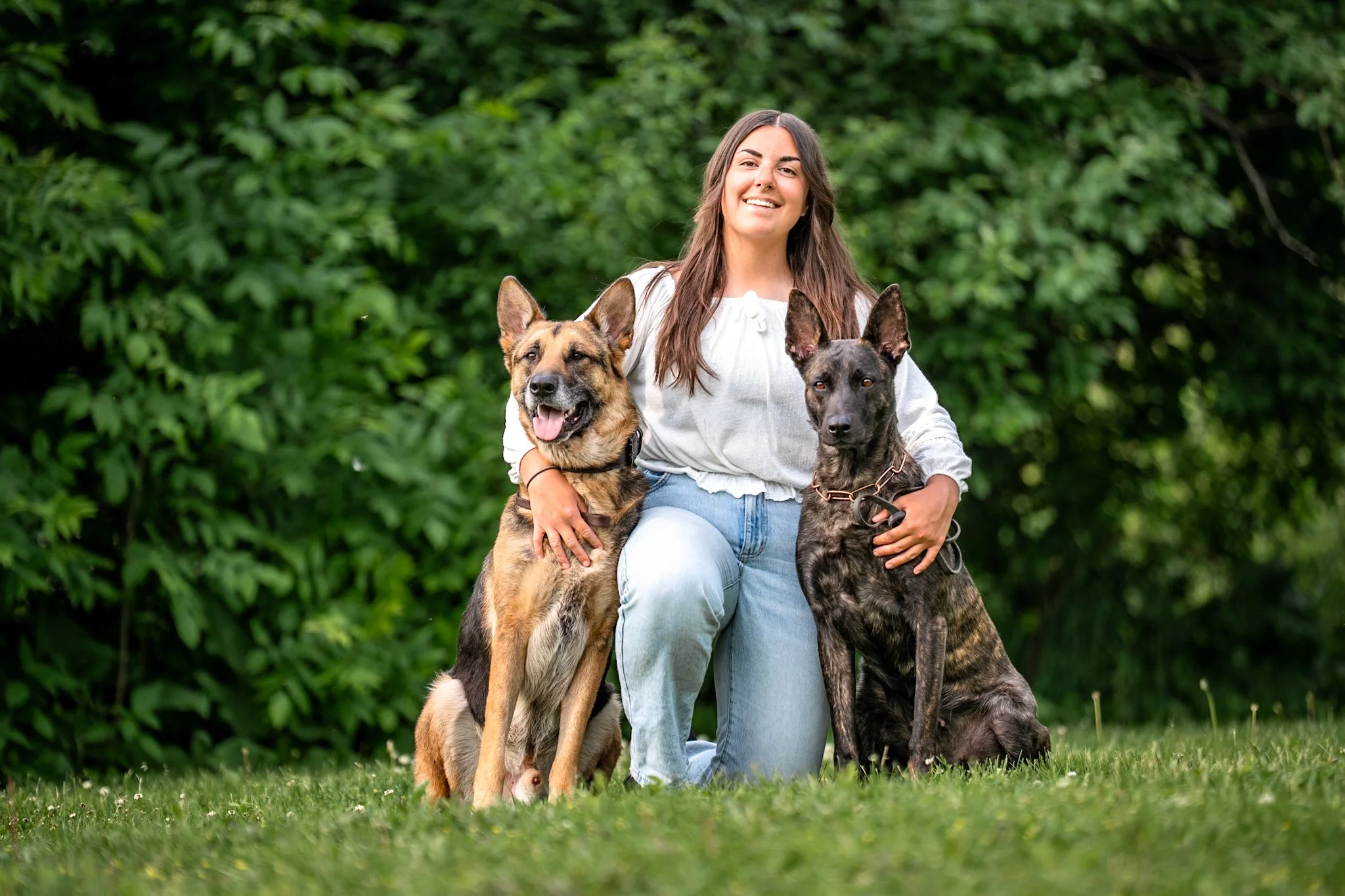 A woman kneeling on the grass outdoors surrounded by two dogs, with a lush green background.
