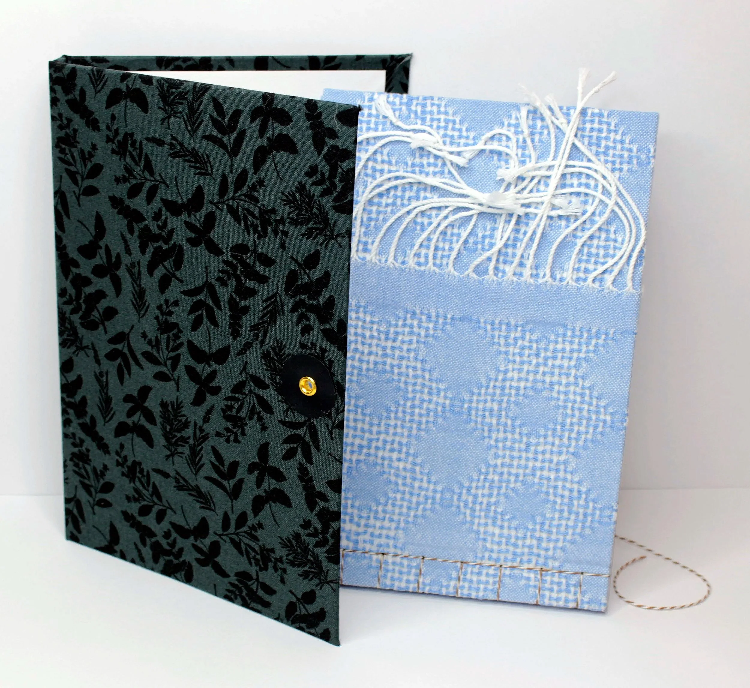 A dark green binder with a leafy pattern partially open, revealing white pages inside, next to a light blue fabric bag with white embroidery and fringe detail.