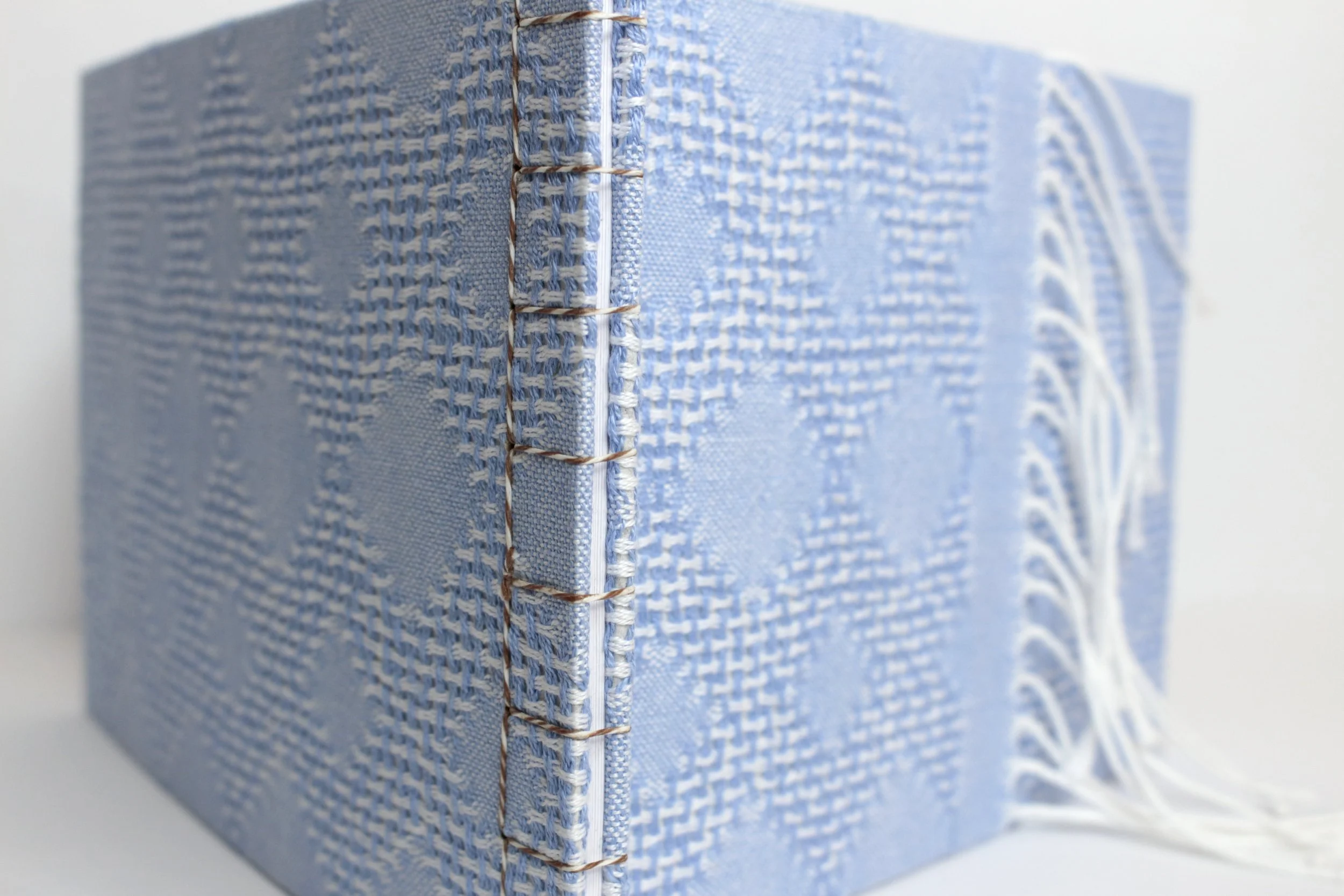 Close-up of a light blue book with textured fabric cover and visible stitching along the spine, lying on a white surface.