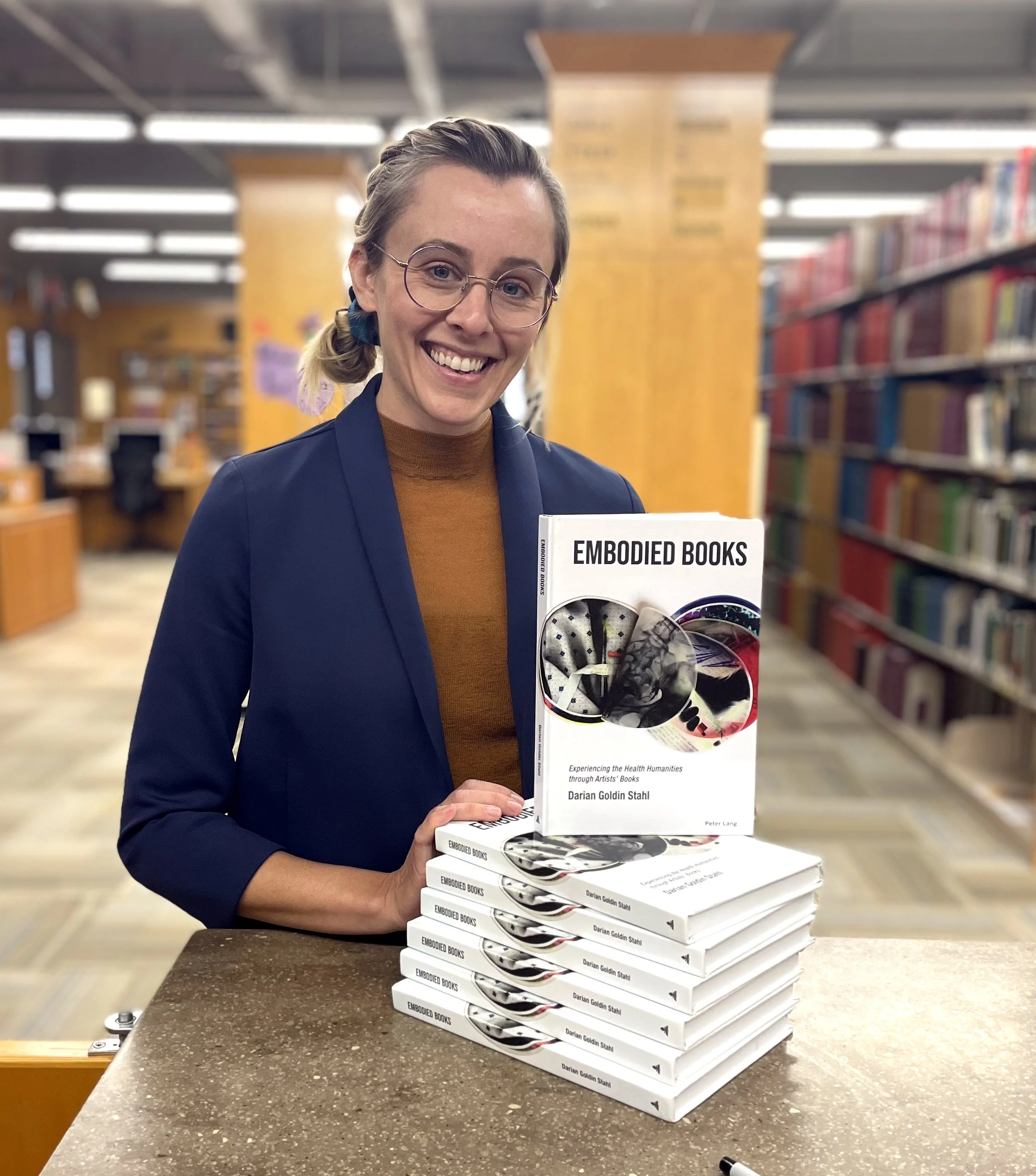 A woman with glasses and a smile holding a book titled 'Embodied Books' in a bookstore, with a stack of similar books on the counter.