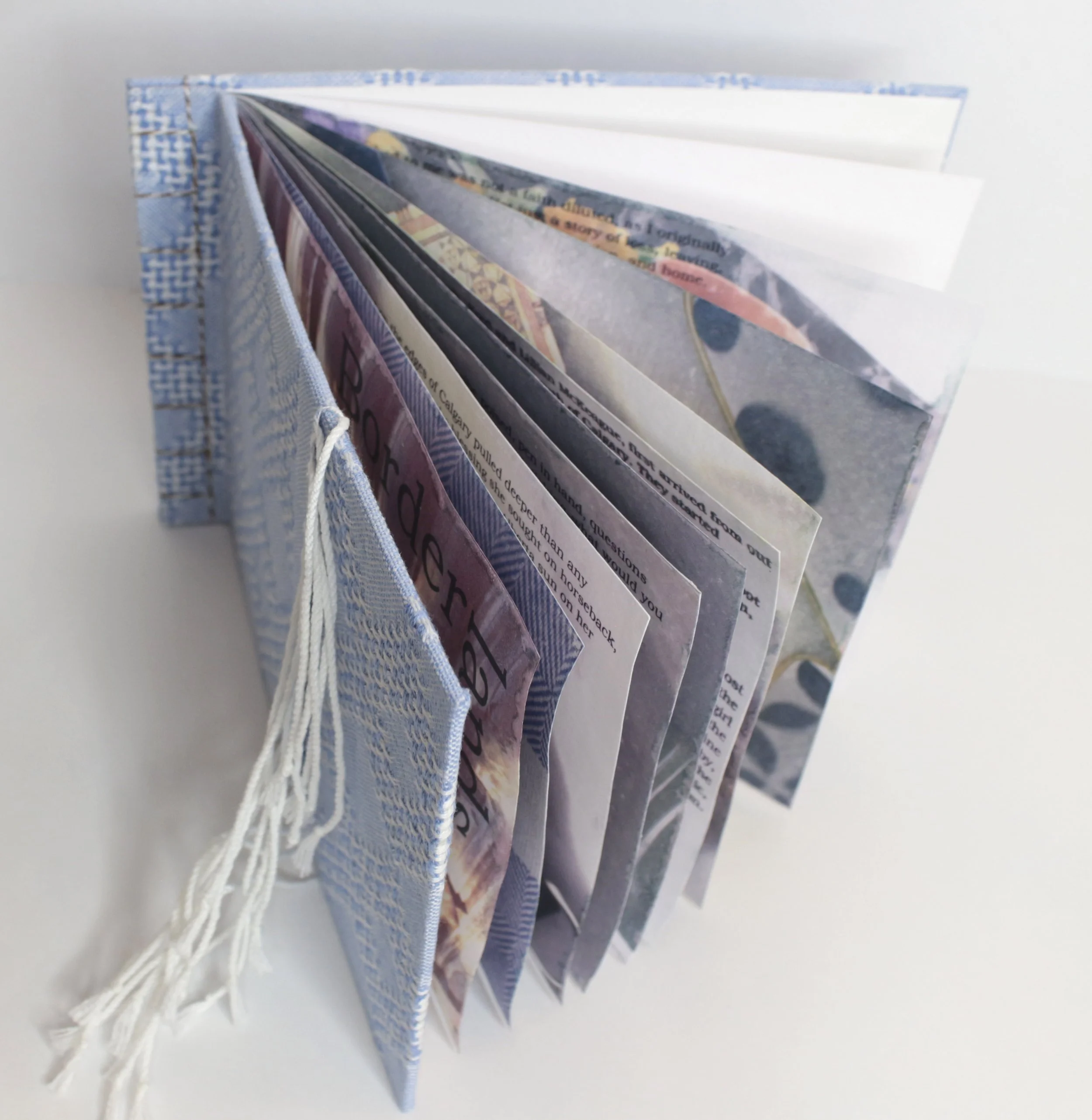 Open handmade paper art journal with fabric cover, displaying layered pages with colorful artwork and text on white background.