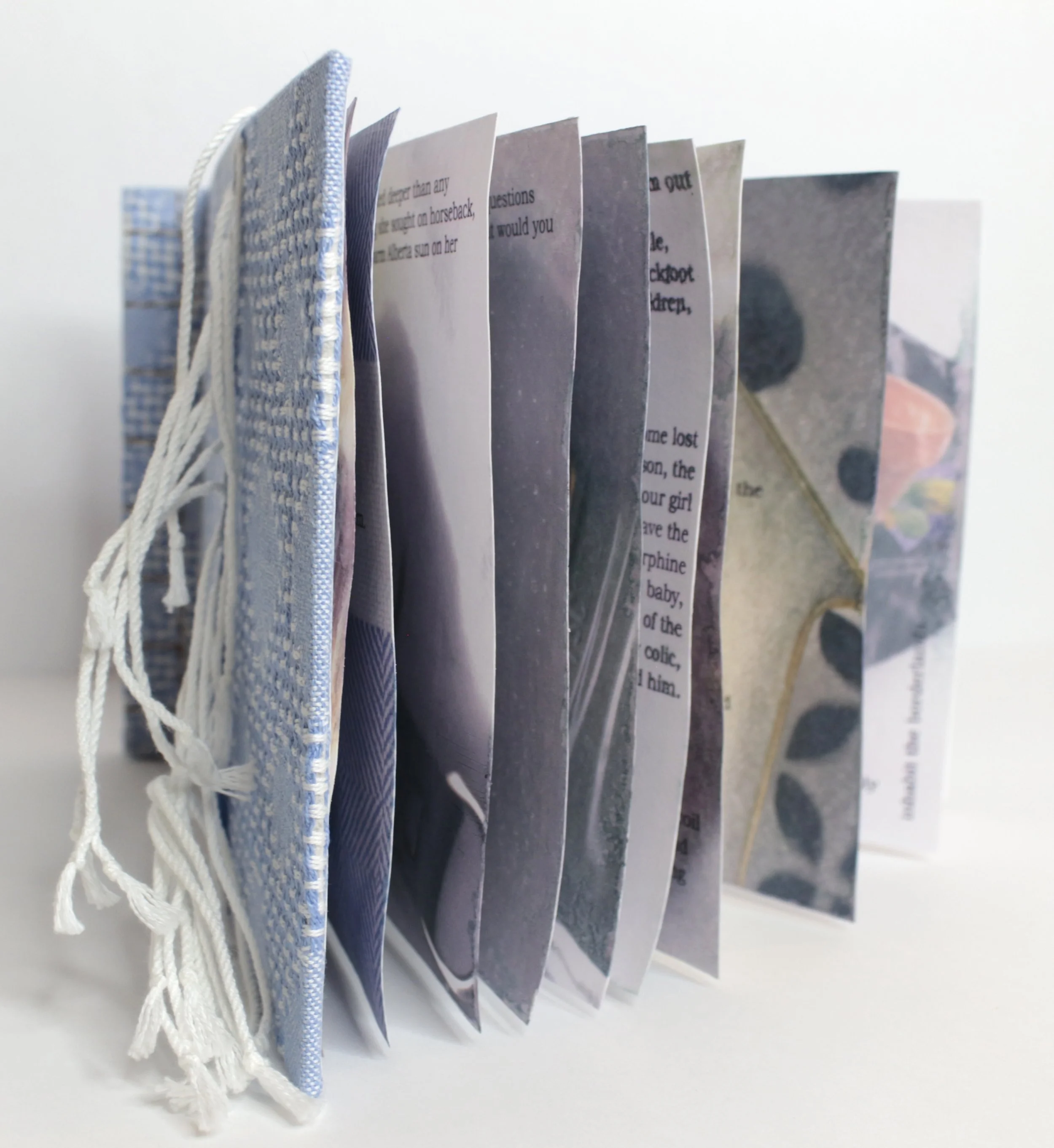 Photo of a handmade book with a blue cloth cover and white tassels on the side, standing open with pages containing printed or handwritten text and images.
