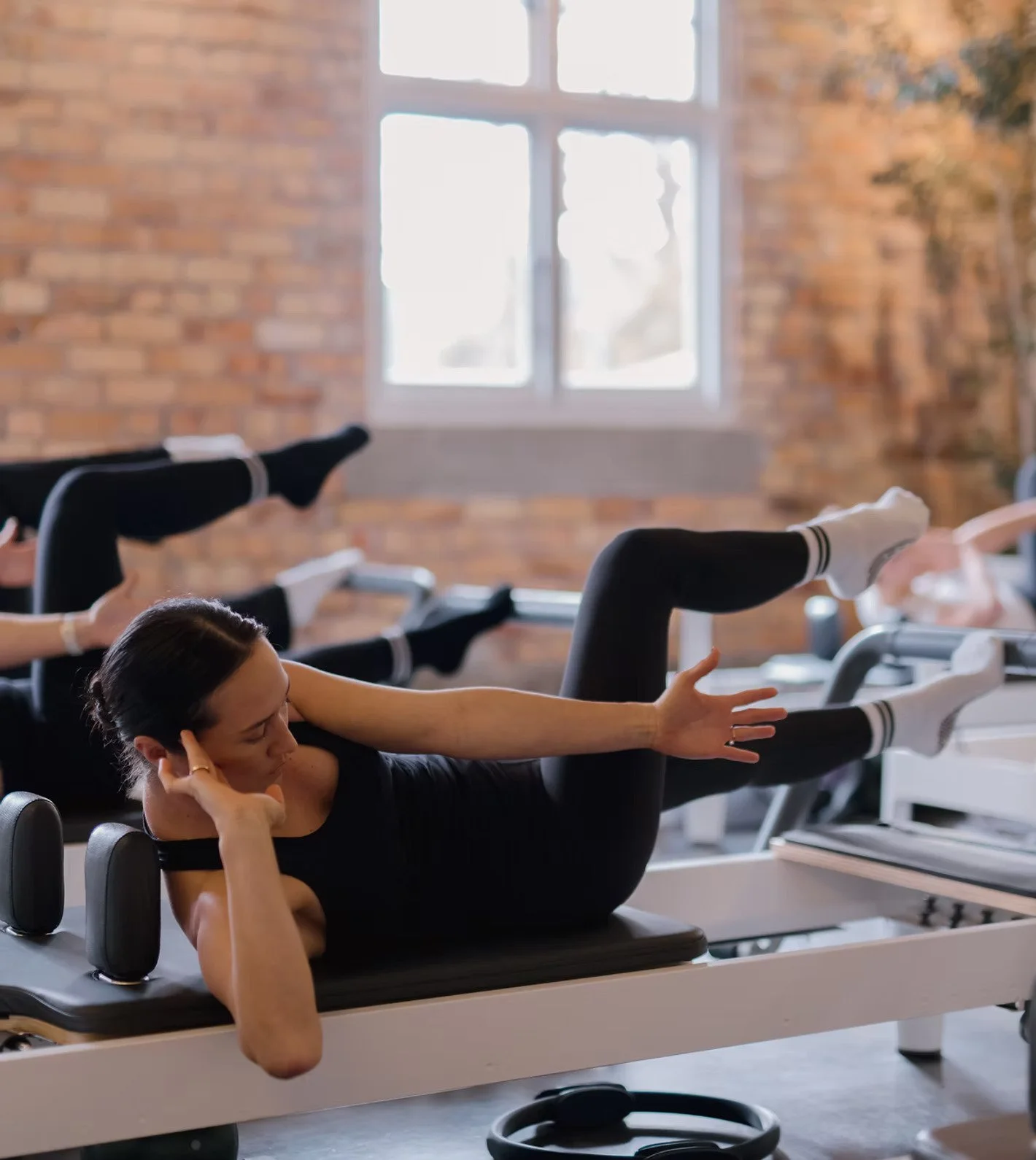 Women doing Pilates exercises on reformer machines in a studio with brick walls and large window.