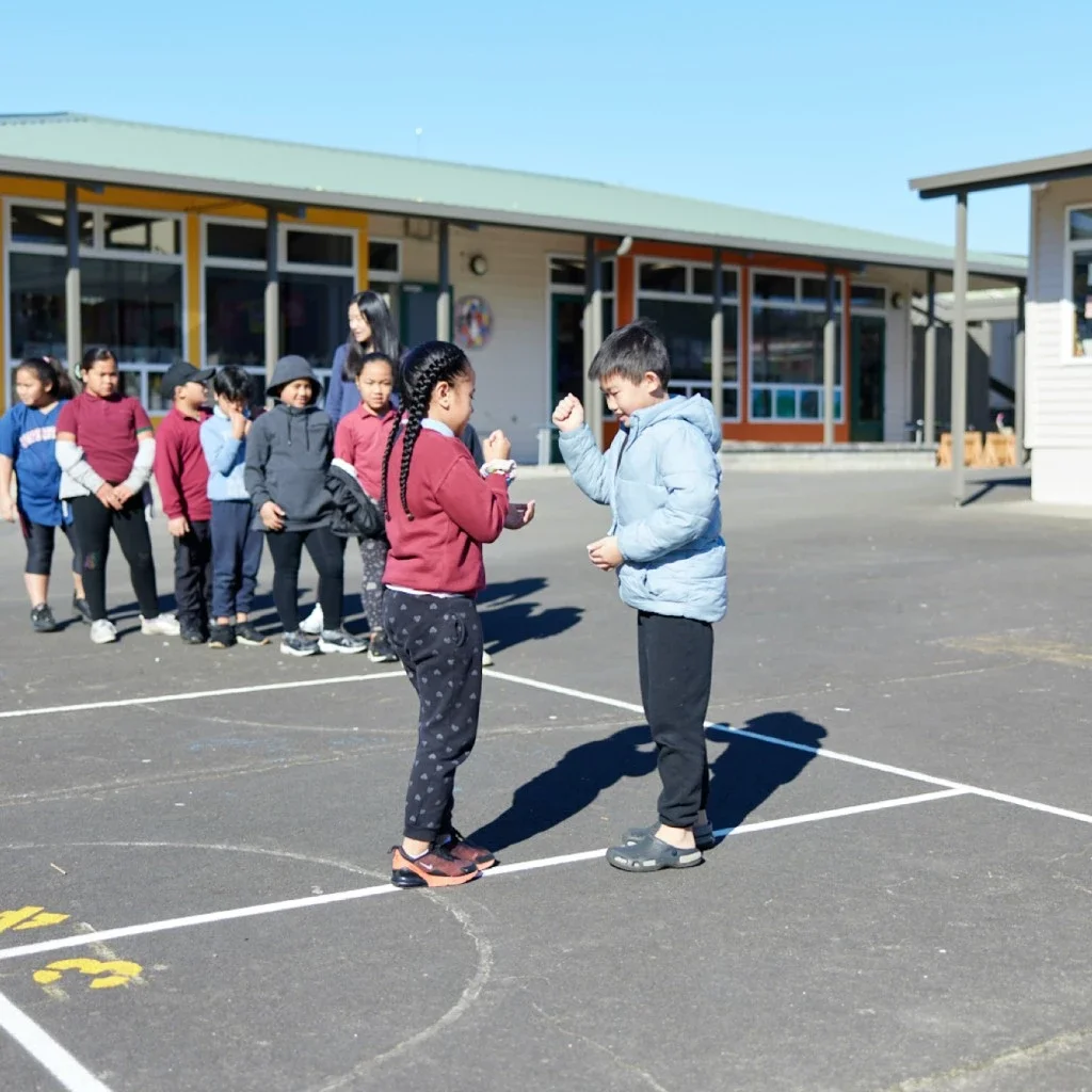 Children standing in line outside school, with two children in front facing each other, appearing to compete or play a game, on a bright sunny day.