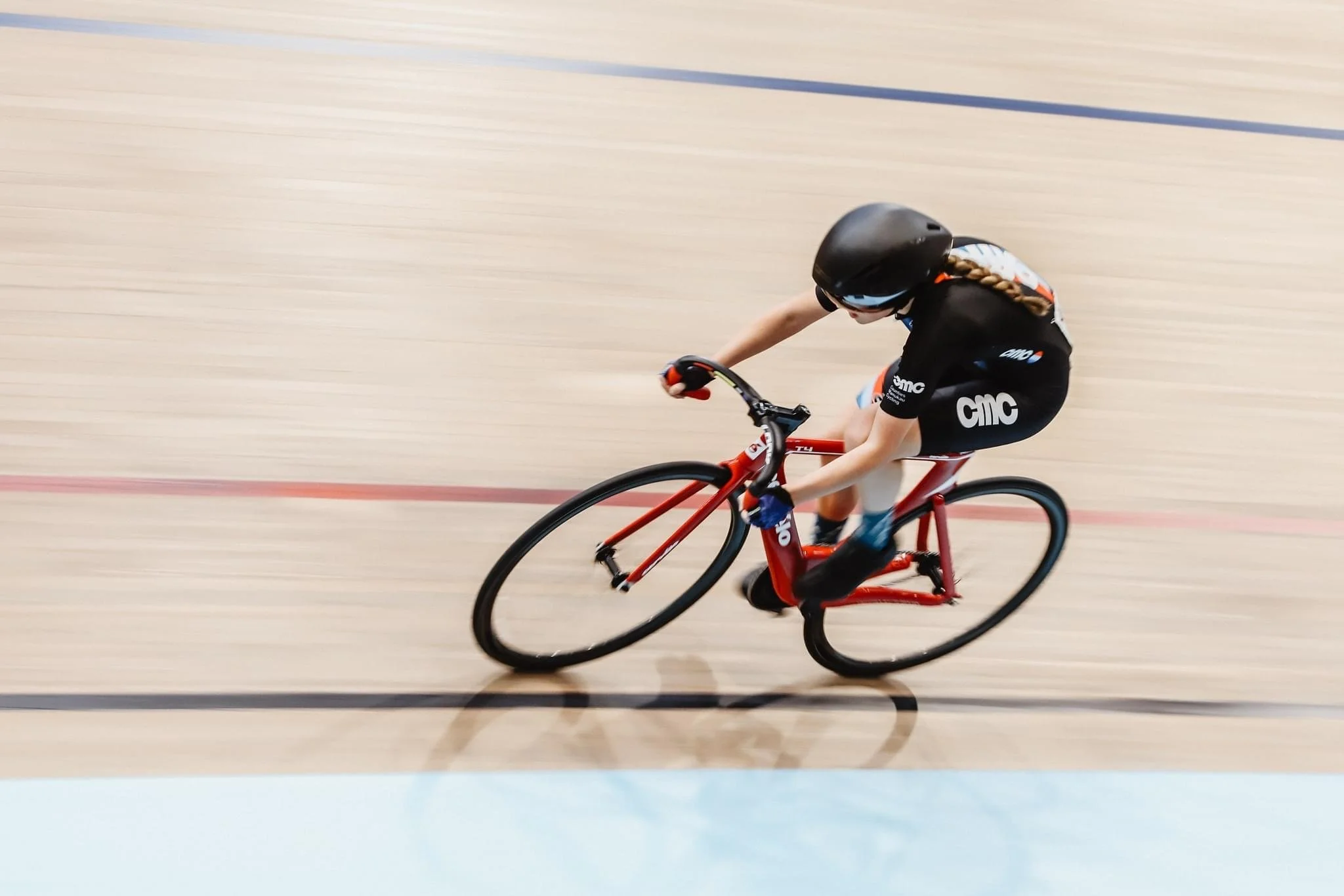 A female cyclist in black and orange attire riding a red bike on an indoor velodrome track.