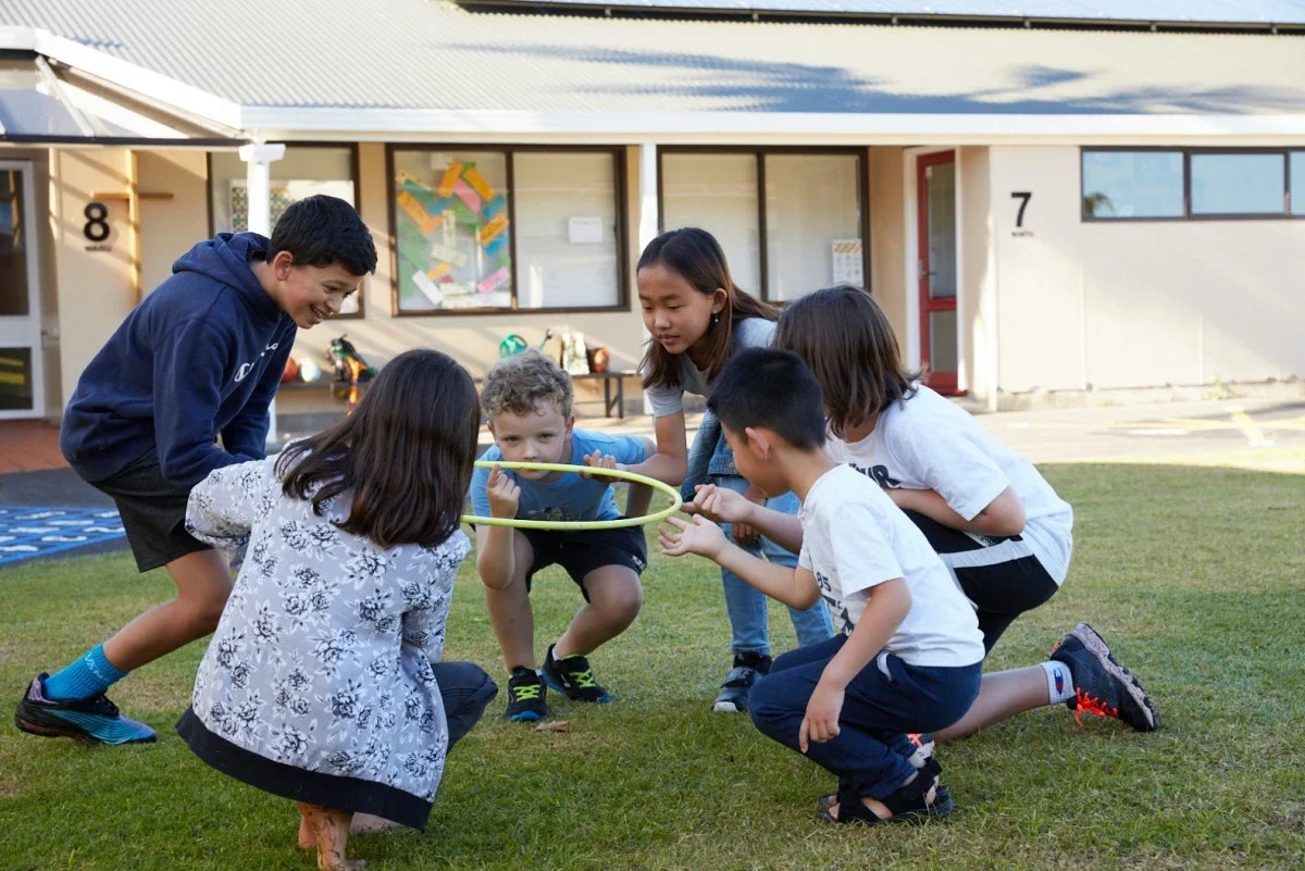 Group of six children playing with a yellow hoop outside on a grassy area in front of a building.