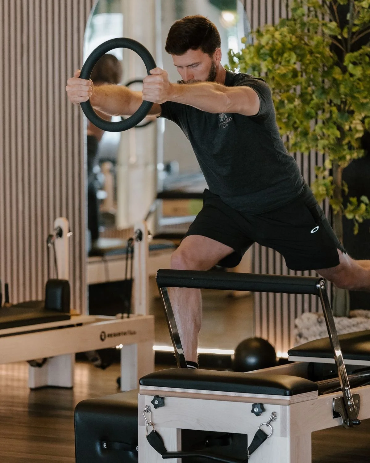 A man performing a workout using a Pilates ring in a gym, standing on a block with one leg while holding the ring with both hands.