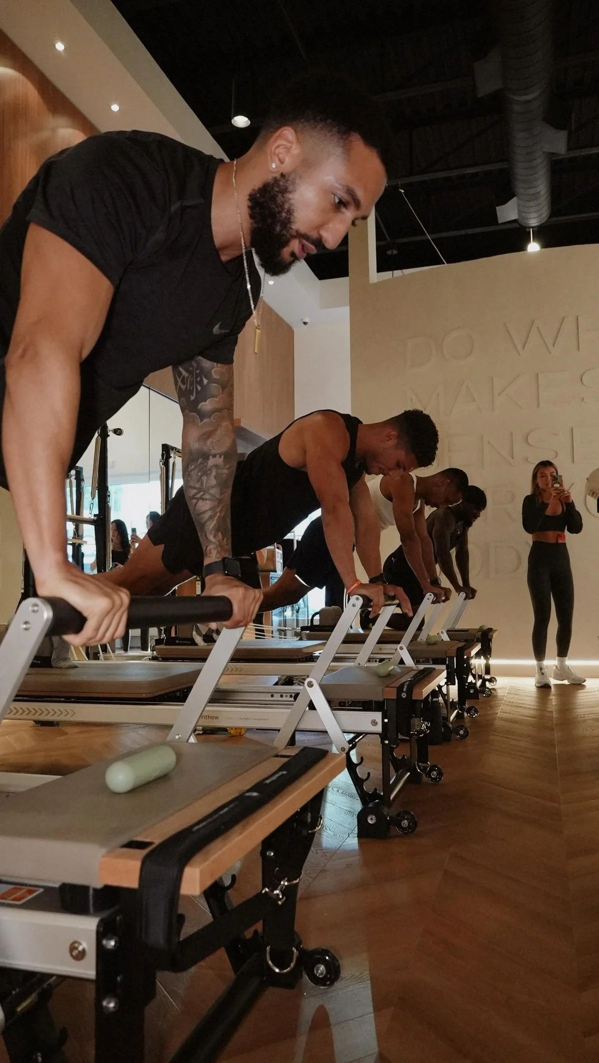 People in black workout clothes doing planks on reformer machines in a fitness studio, with a woman taking a picture in the background.