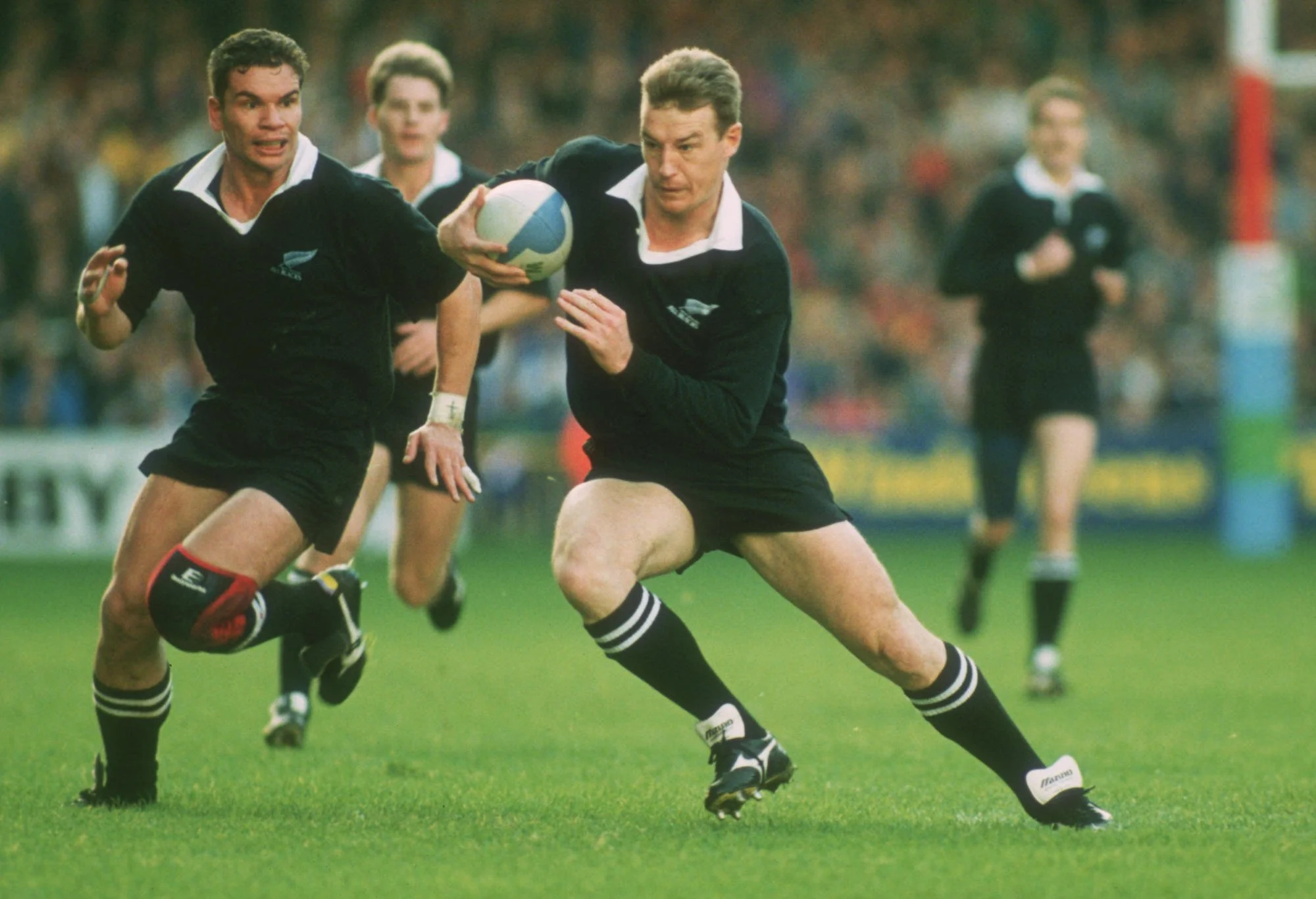 Two rugby players in black uniforms competing for the ball on a green field, with other players and spectators in the background.