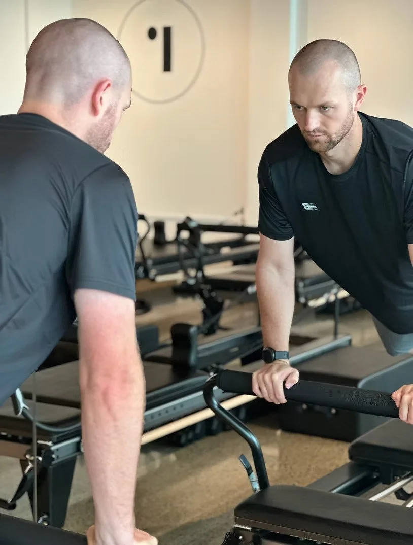 Two men with shaved heads and athletic builds performing a push-up exercise on parallel bars in a gym, with Pilates reformer equipment in the background.