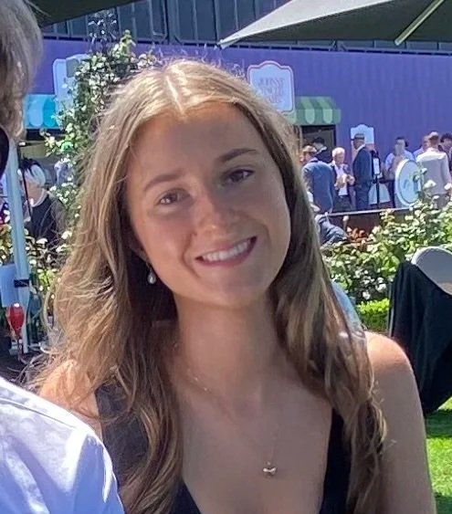 A young woman with long light brown hair smiling outdoors at a sunny event.