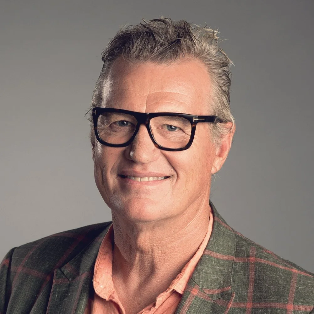 A man with gray hair and glasses wearing a plaid jacket and a peach shirt, smiling against a gray background.