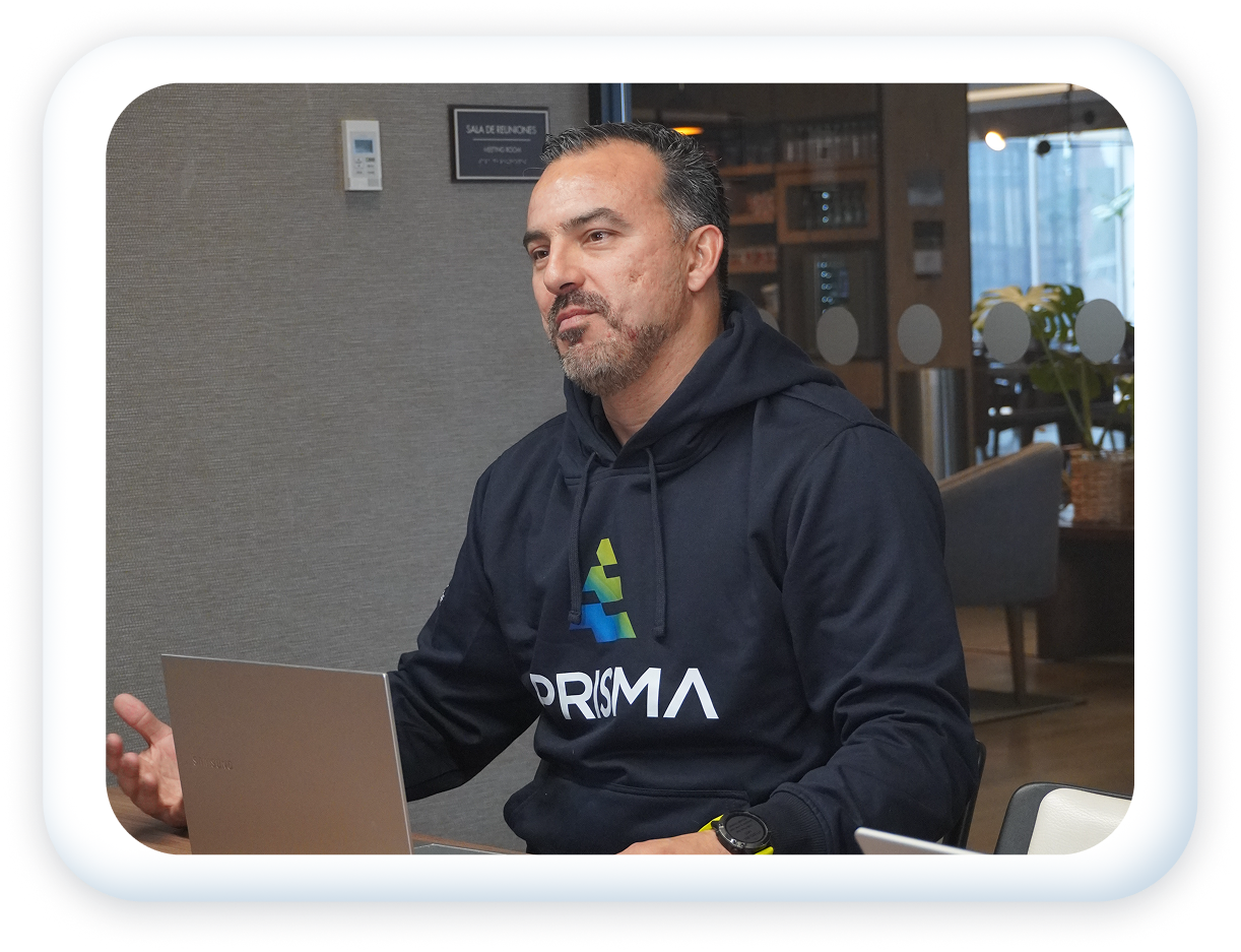 A man wearing a black PRISMA hoodie is sitting at a table with a laptop in front of him, gesturing with his right hand, inside a modern office or conference room.