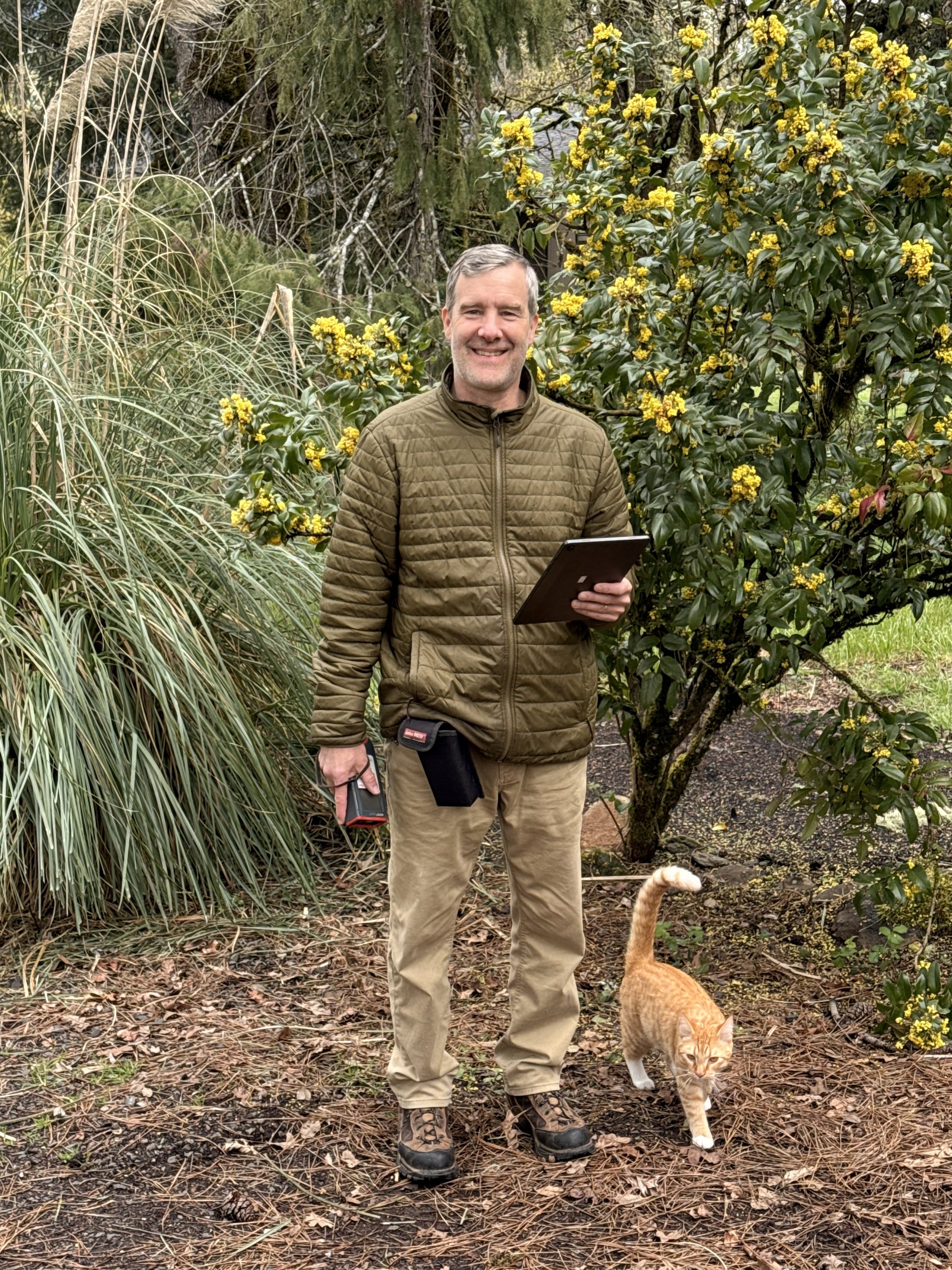 A smiling man in outdoor clothing standing in a garden with yellow flowering bushes and tall grasses, holding a tablet and a device, with an orange cat nearby.
