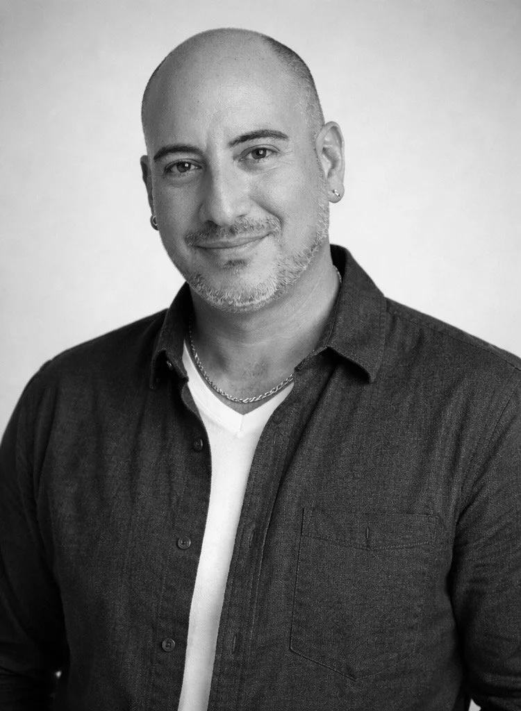 Black and white portrait of a man with a shaved head, beard, and earrings, wearing a dark shirt over a white T-shirt, smiling slightly.