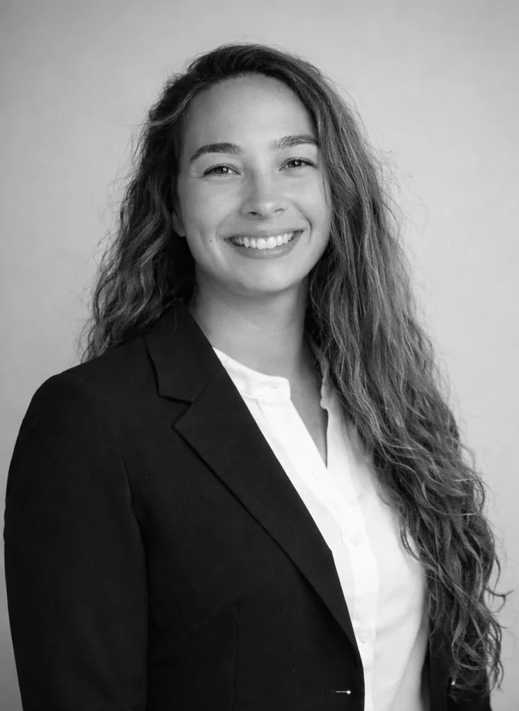Black and white professional portrait of a young woman with long, curly hair, smiling, wearing a dark blazer over a light shirt, against a plain background.