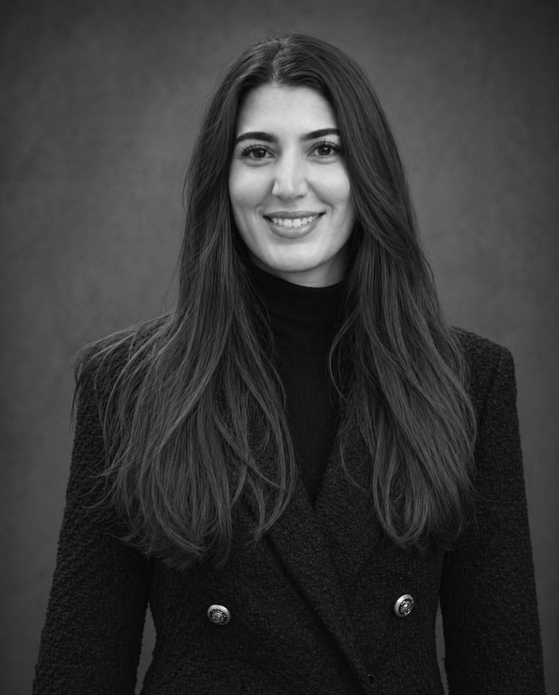 Black and white portrait of a woman with long hair, smiling, wearing a dark blazer and turtleneck, standing against a plain background.