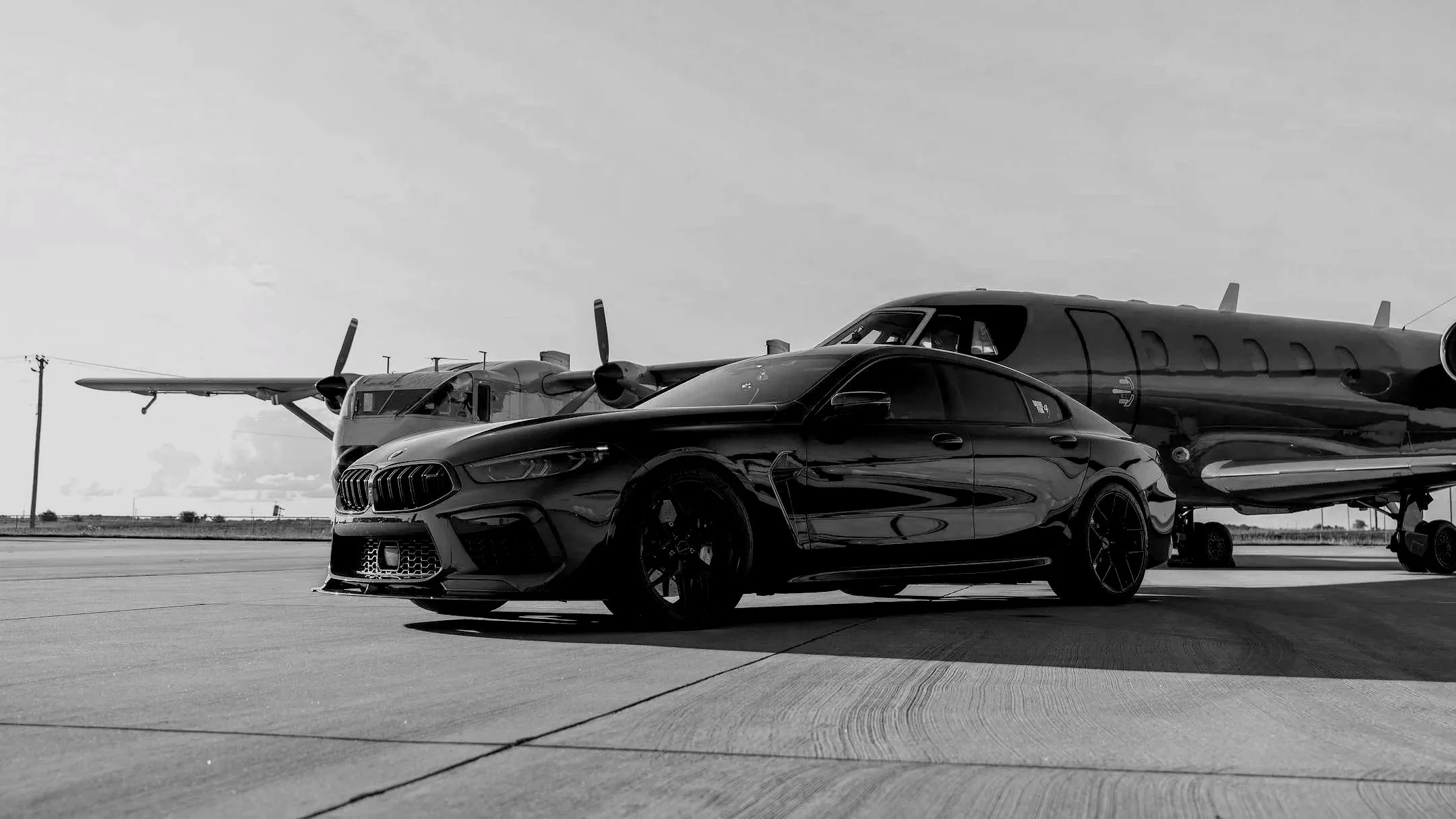 Black sports car parked on tarmac in front of a small propeller aircraft at an airfield on a cloudy day, black and white photo.