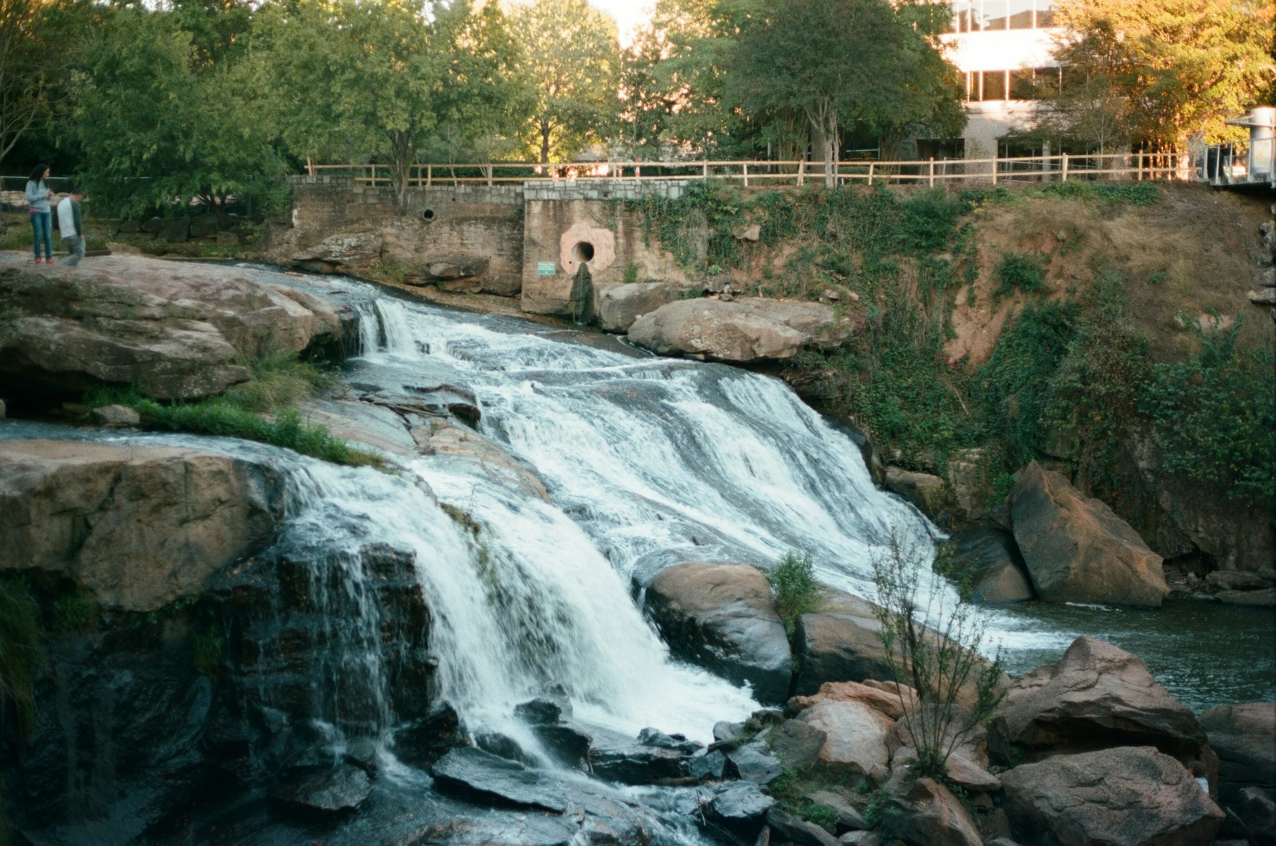 A small waterfall cascading over rocks with two people standing on a rocky outcrop to the left, trees and a building in the background.