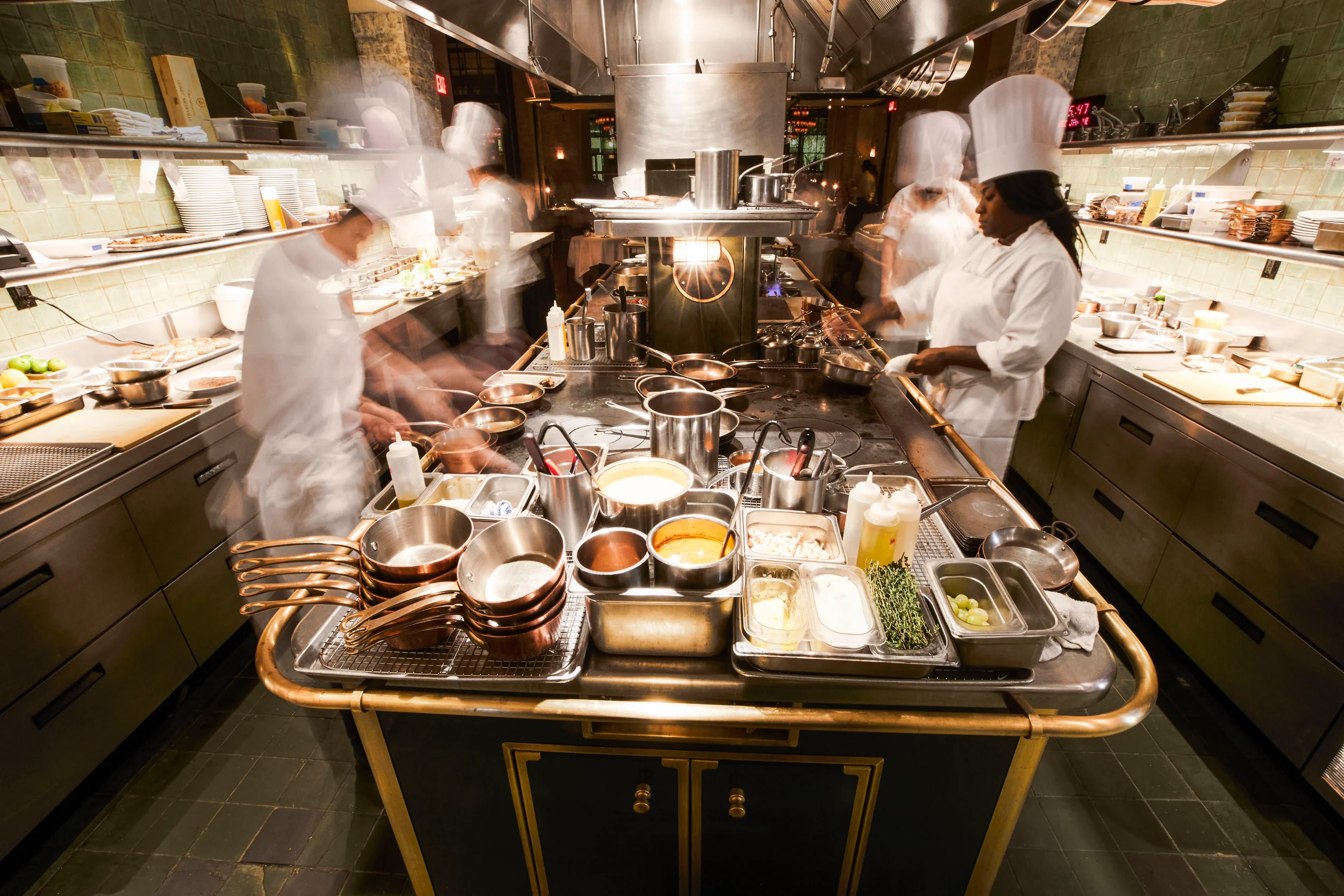 Kitchen with multiple chefs in white uniforms and tall hats preparing food. The counter is filled with pots, pans, utensils, and ingredients. The scene shows motion blur, indicating active cooking.