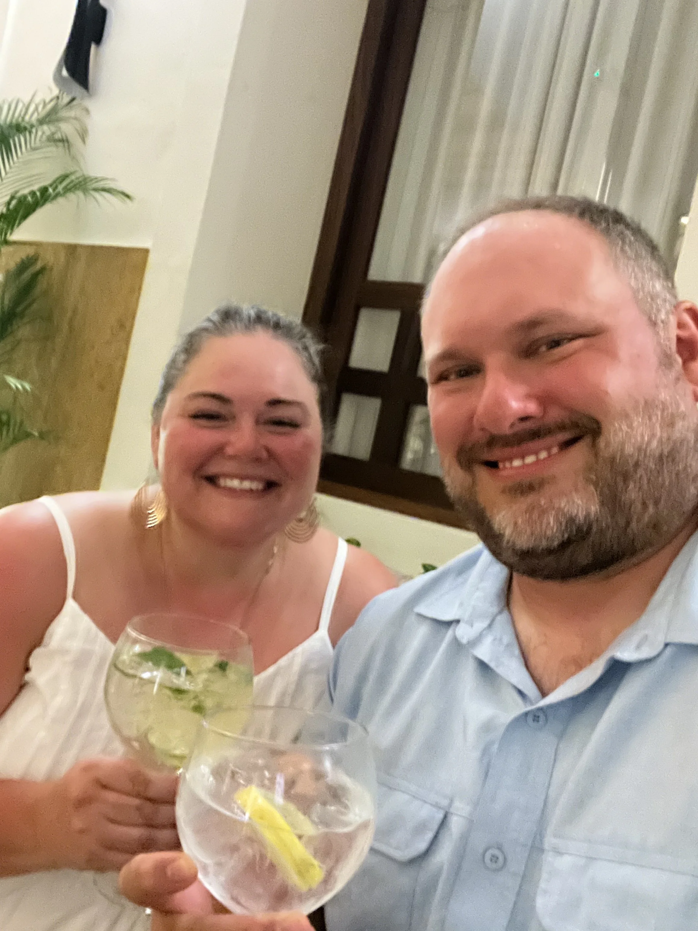 Two smiling people, a woman and a man, holding drinks, in a cozy indoor setting with plants and a window in the background.