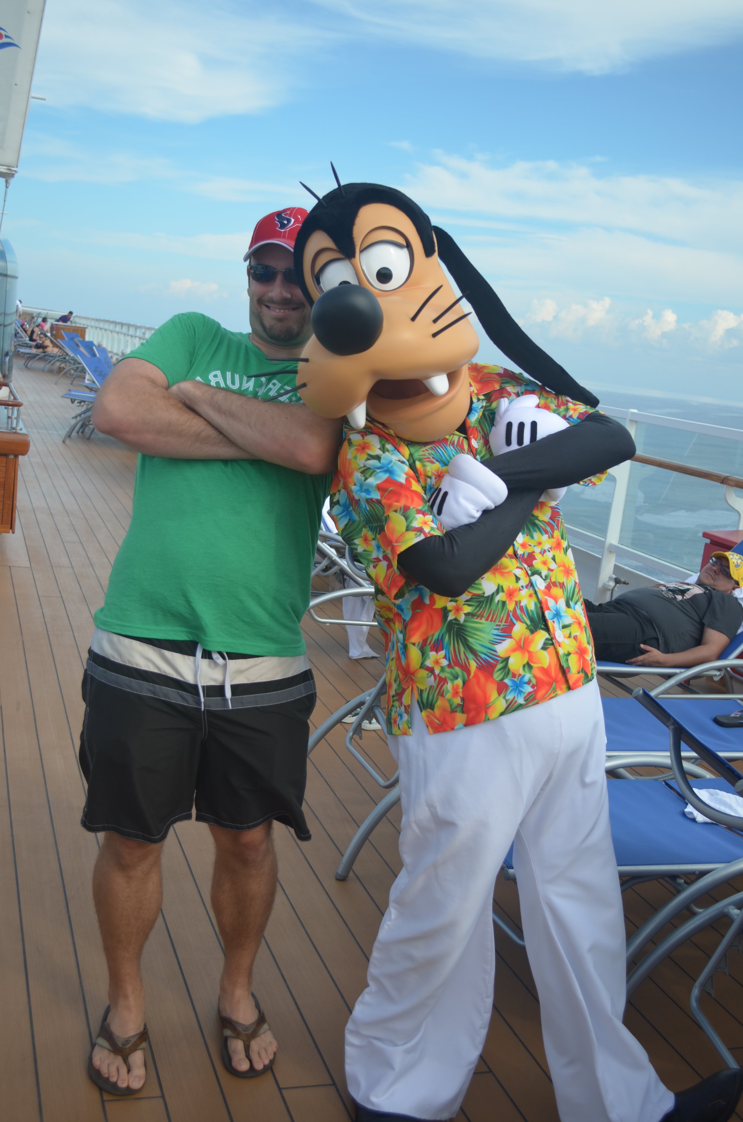 A man in a green T-shirt, black shorts, and flip-flops standing with his arms crossed on a cruise ship deck, posing with a person in a Goofy costume wearing a colorful Hawaiian shirt and white pants.