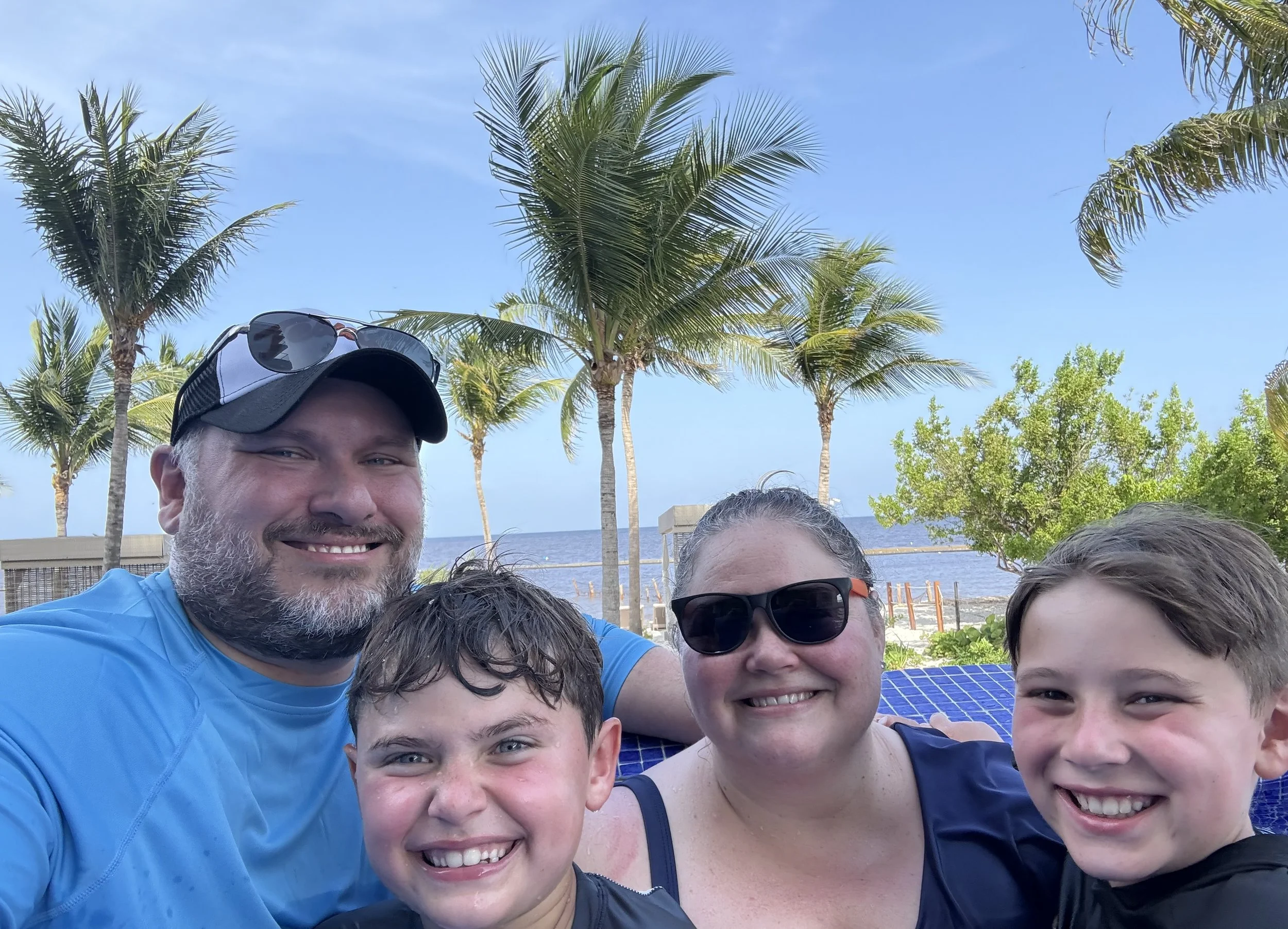 A family of four smiling for a selfie at the beach with palm trees and ocean in the background.