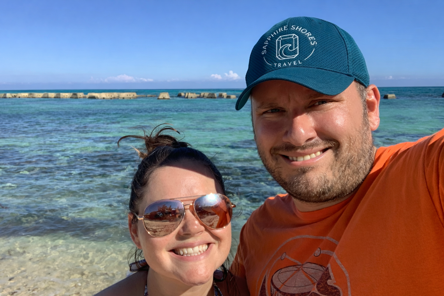 Smiling couple taking a selfie on a beach with clear blue water and a blue sky in the background.