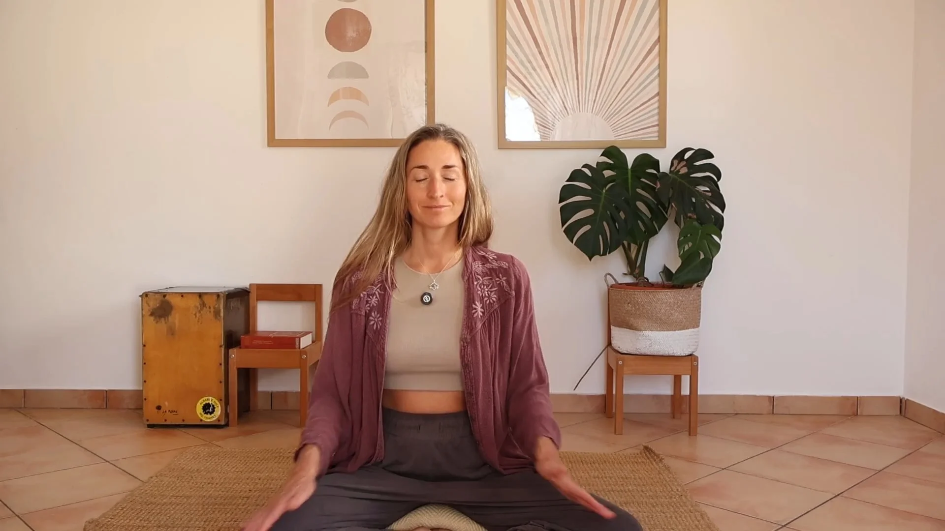 A woman practicing meditation or yoga indoors, sitting cross-legged on a cushion with her eyes closed and hands resting on her knees. The room has minimalist decor with framed abstract artwork on the wall, a large leafy potted plant, and a wooden floor.