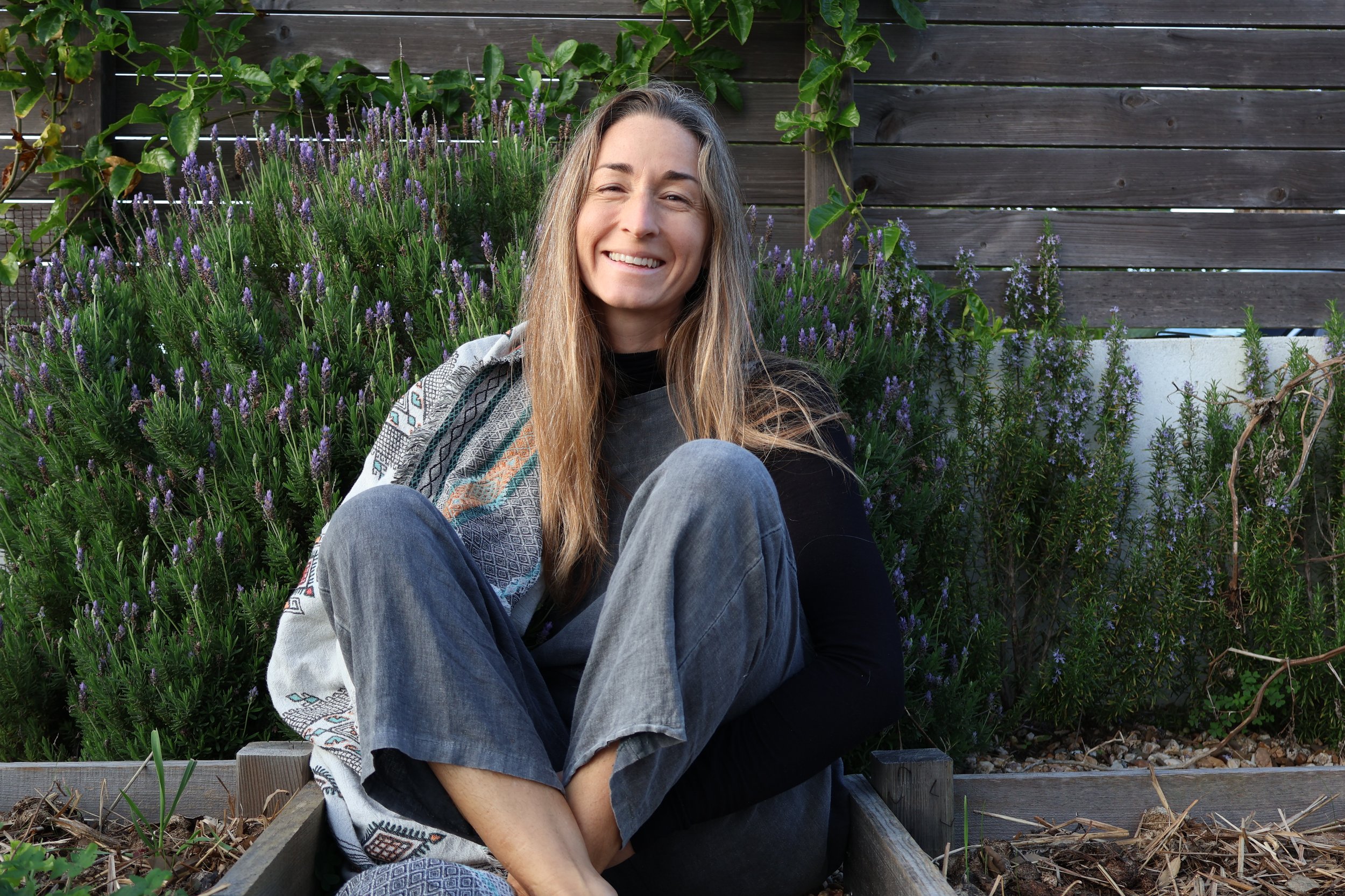 A smiling woman with long hair sitting cross-legged in a garden with purple flowering plants and a wooden fence background.