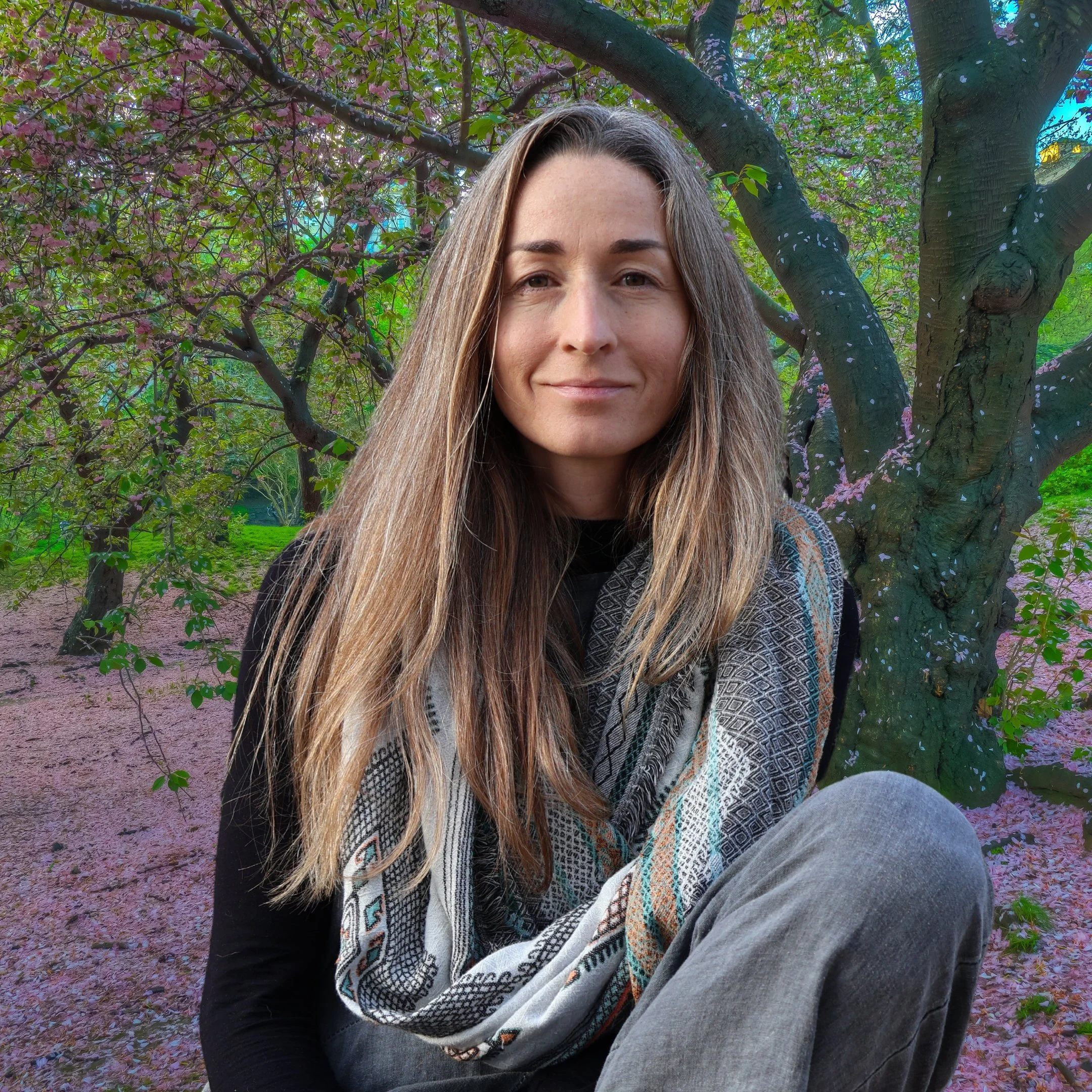 A woman with long light brown hair sitting outdoors near a tree with purple blossoms, wearing a patterned scarf and a black top.