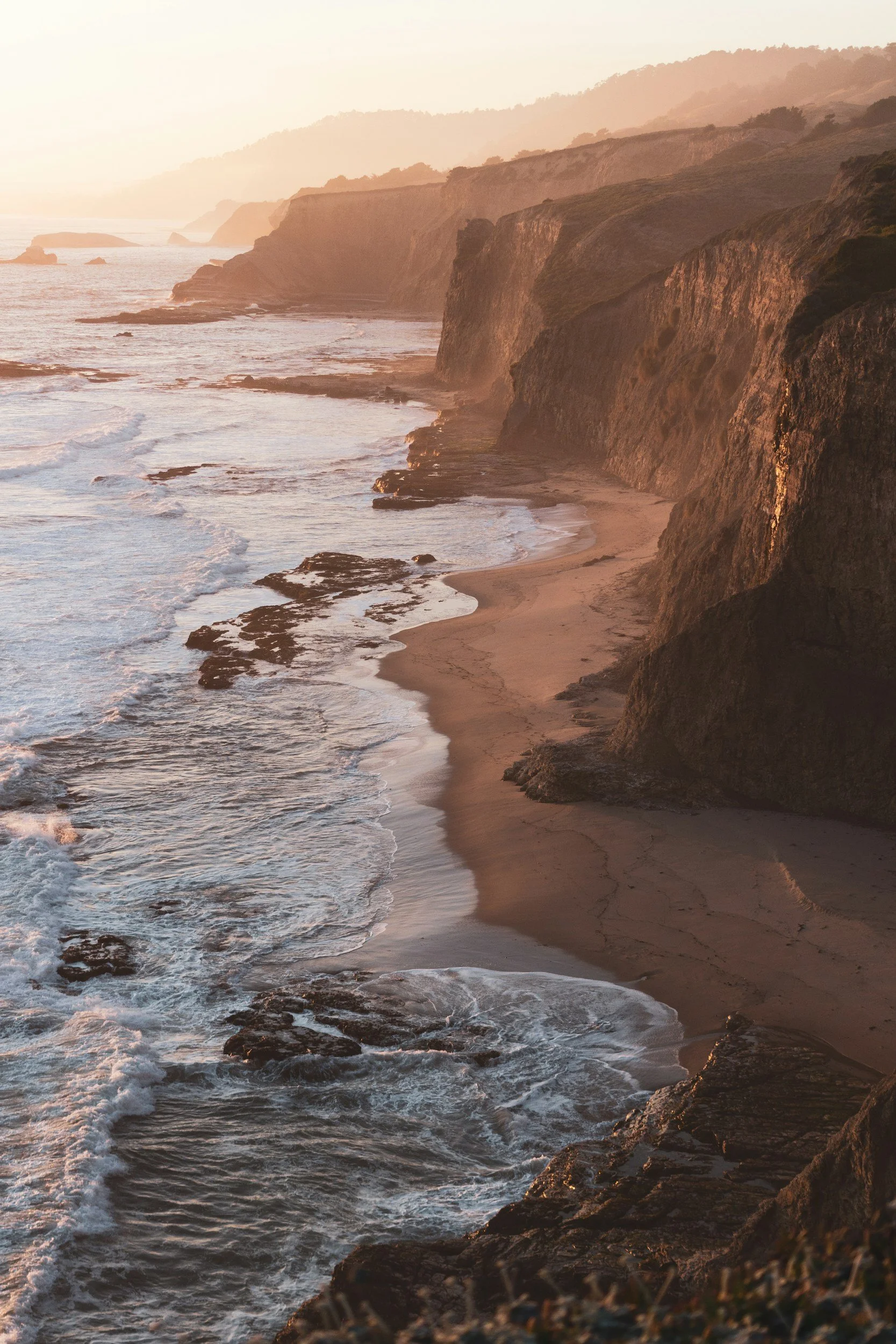 Cliffs and coastline along the ocean during sunset with waves crashing on the shore.