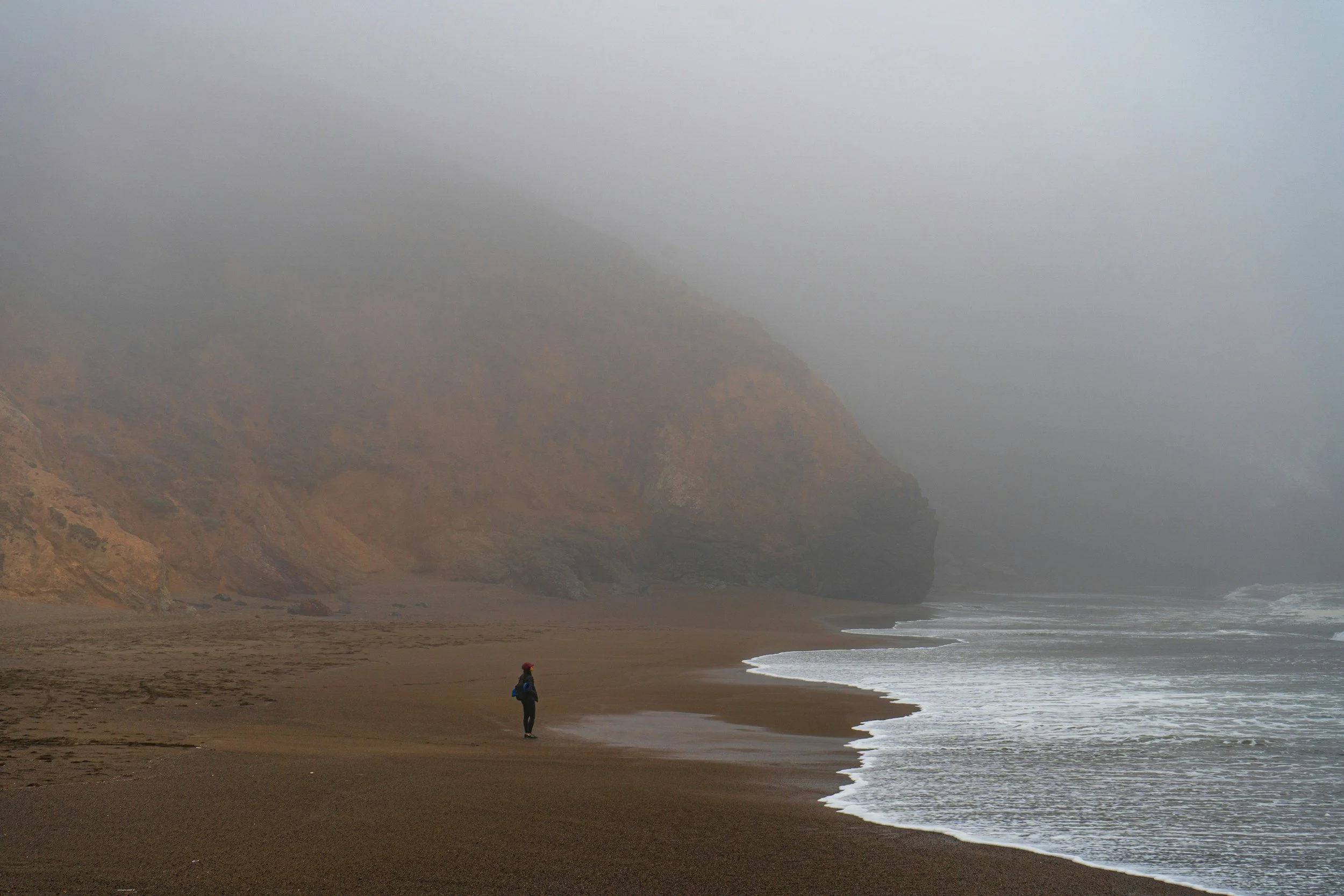 A person standing alone on a foggy beach, looking at the ocean waves, with large cliffs in the background shrouded in mist.