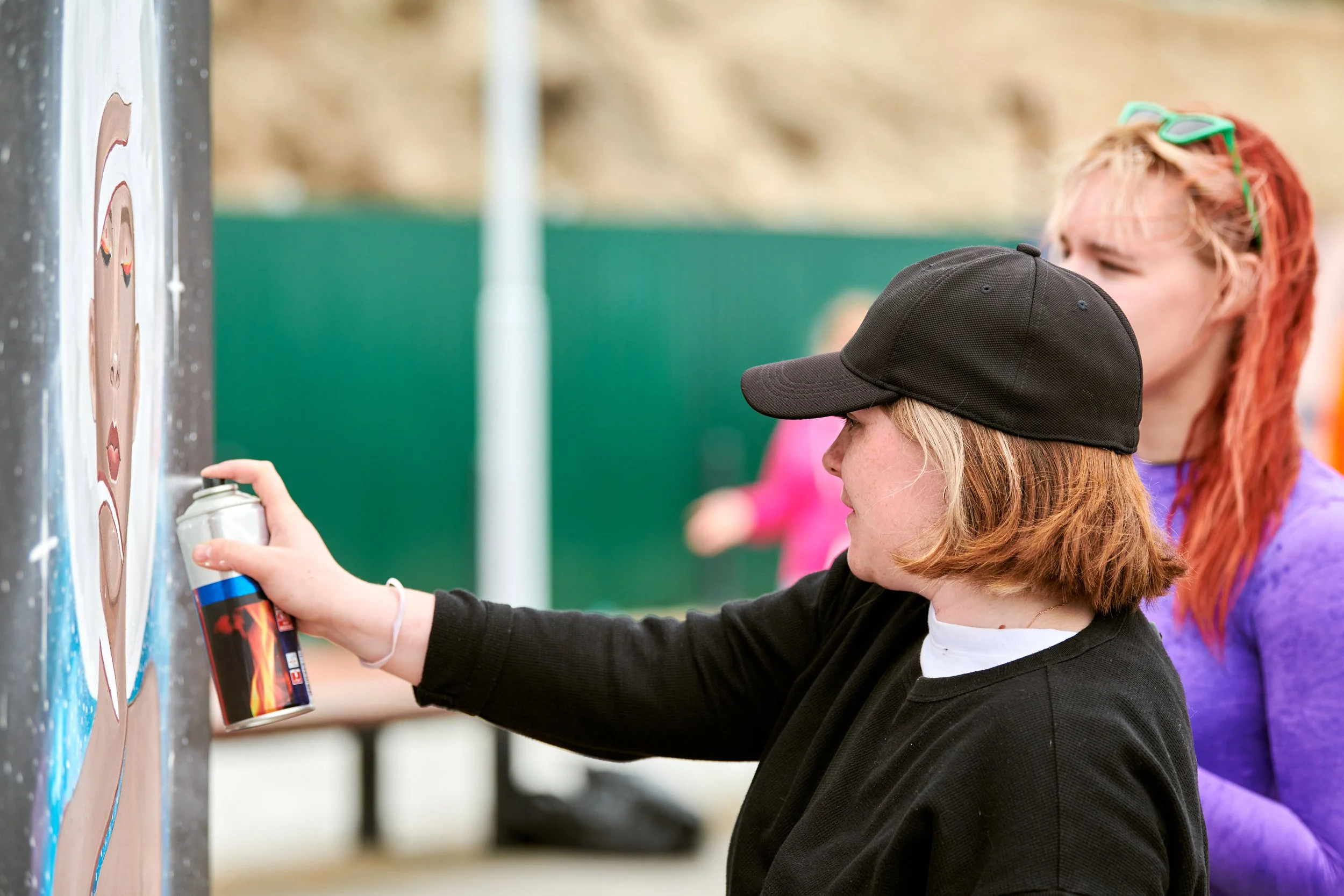 A young person wearing a black baseball cap and black sweatshirt is spray-painting a graffiti on a wall outdoors. Behind them, another person with red hair, sunglasses on their head, and a purple shirt watches.