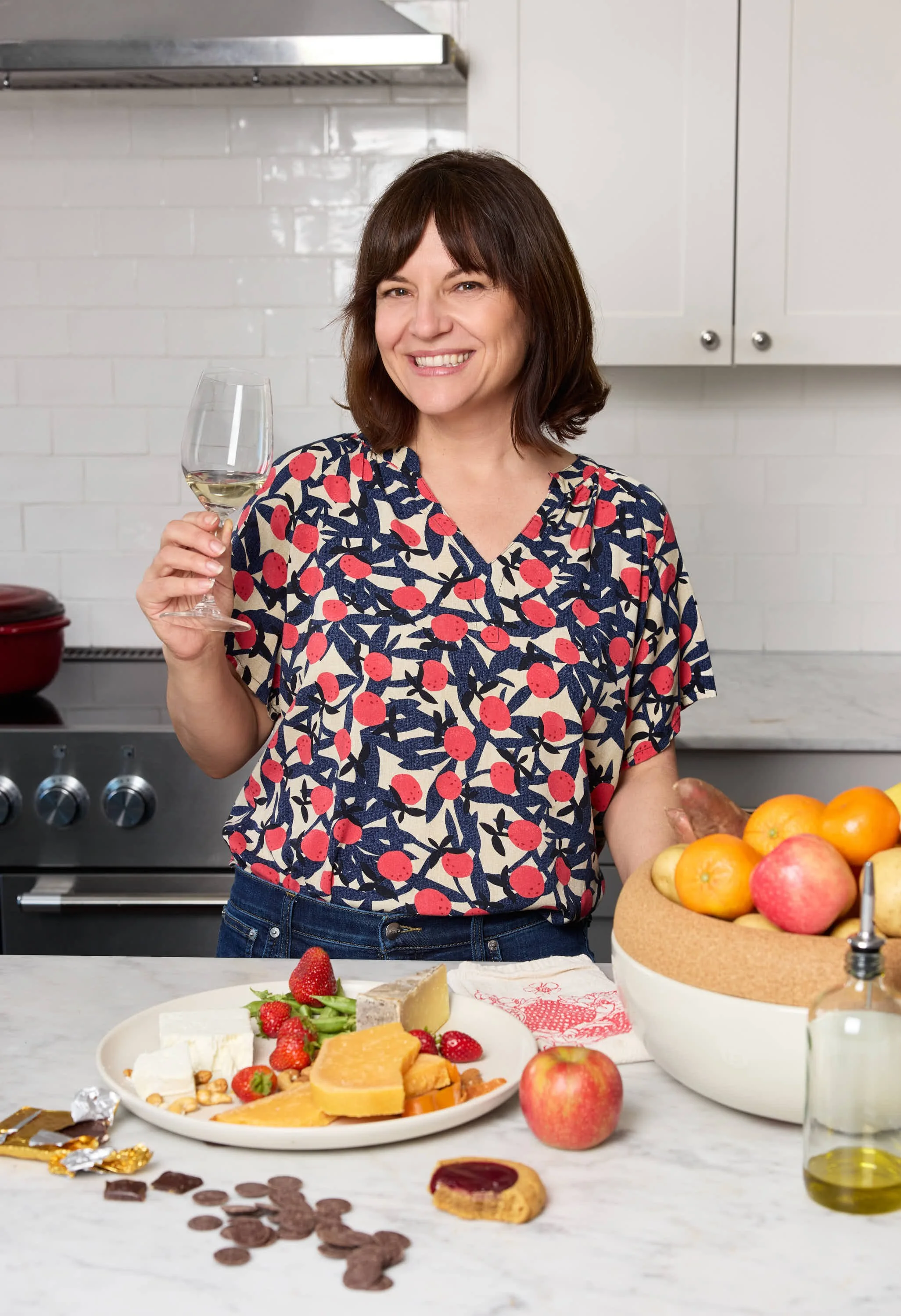 Woman smiling, holding a glass of white wine in a kitchen with a white countertop, bowl of oranges and apples, and a plate of cheese, strawberries, and chocolate chocolates.