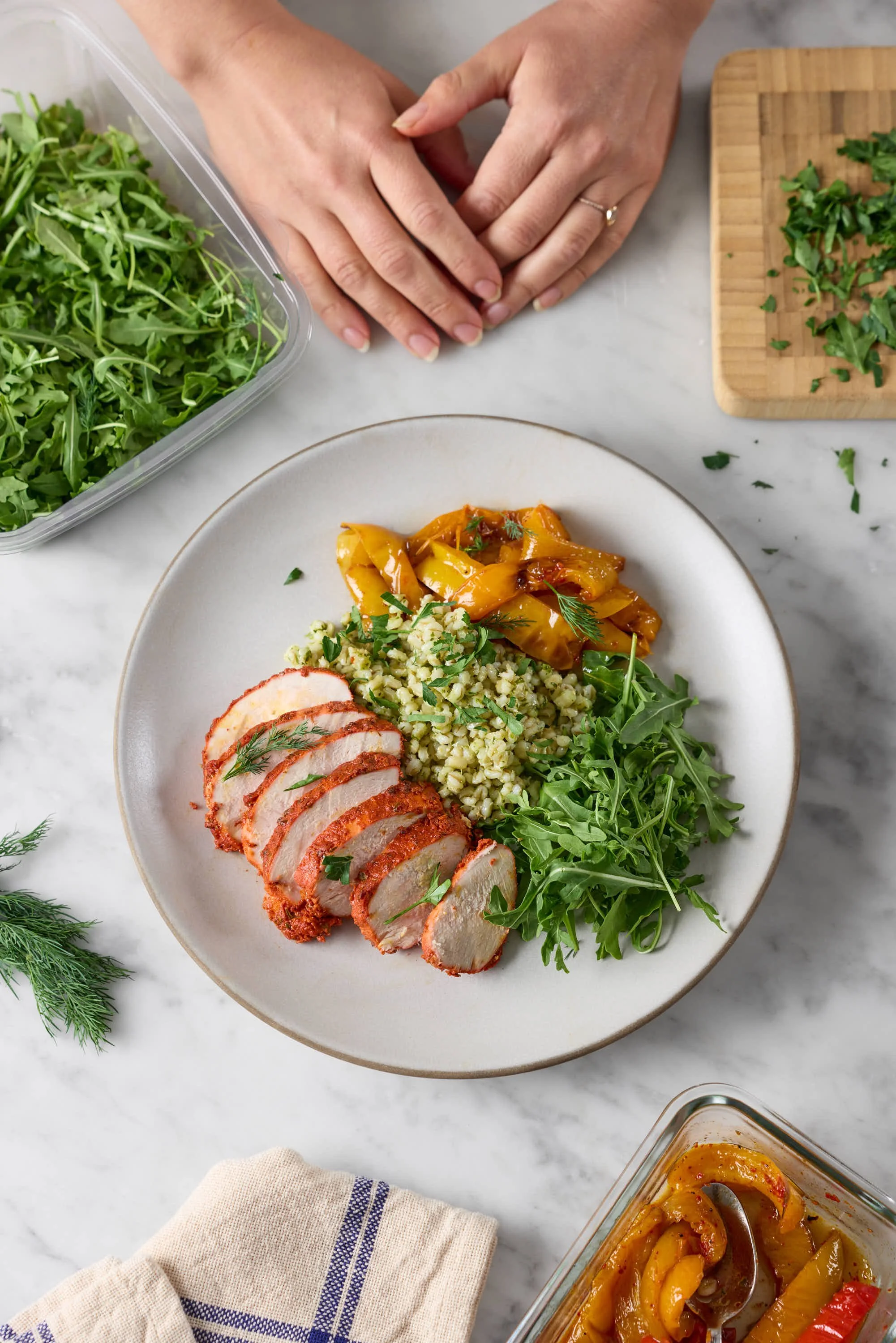 Sliced cooked chicken breast with spices served with grains and roasted yellow bell peppers on a white plate, garnished with herbs, with a side of chopped greens, and a container of roasted peppers nearby, on a marble surface.