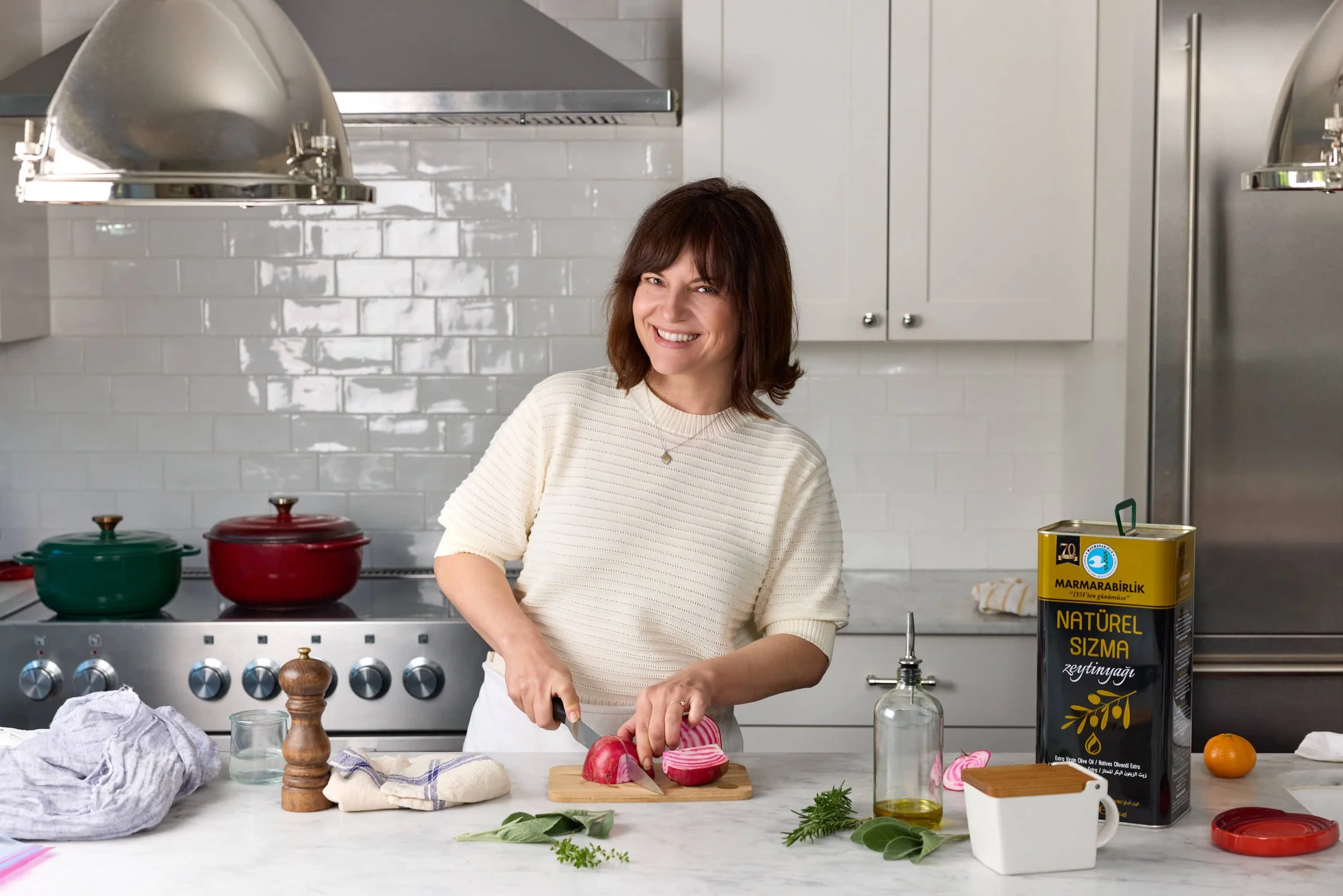 A woman in a cream sweater is slicing an onion at a kitchen counter. The kitchen has a white subway tile backsplash, stainless steel appliances, and various cooking pots and ingredients on the counter, including a can of natural almond paste and fresh herbs.