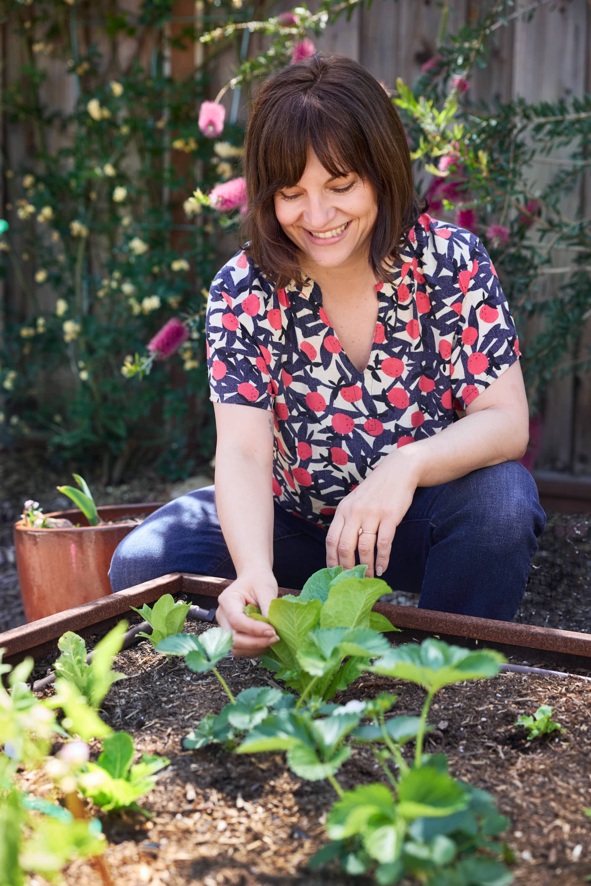 A woman gardening in her backyard, planting or inspecting green leafy plants in a raised garden bed while smiling and enjoying her time outdoors.