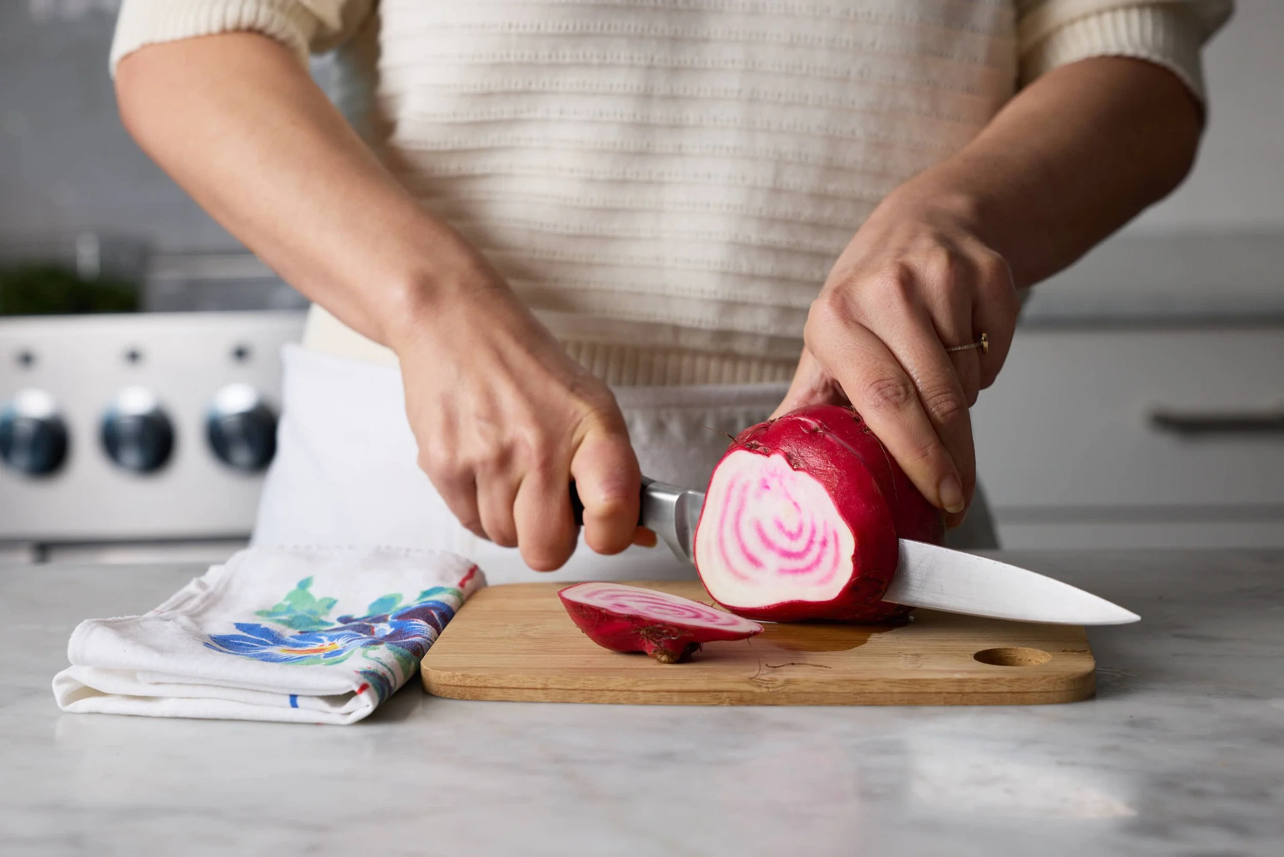 A person slicing a red and white radish on a wooden cutting board in a kitchen, with a colorful cloth and a white napkin nearby.